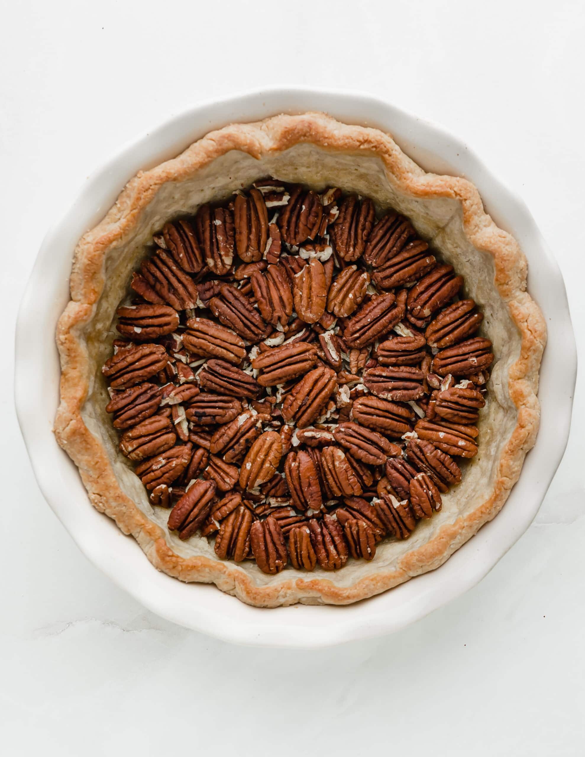 A picture of a white ceramic pie dish, with a par-baked pie crust and toasted pecans.
