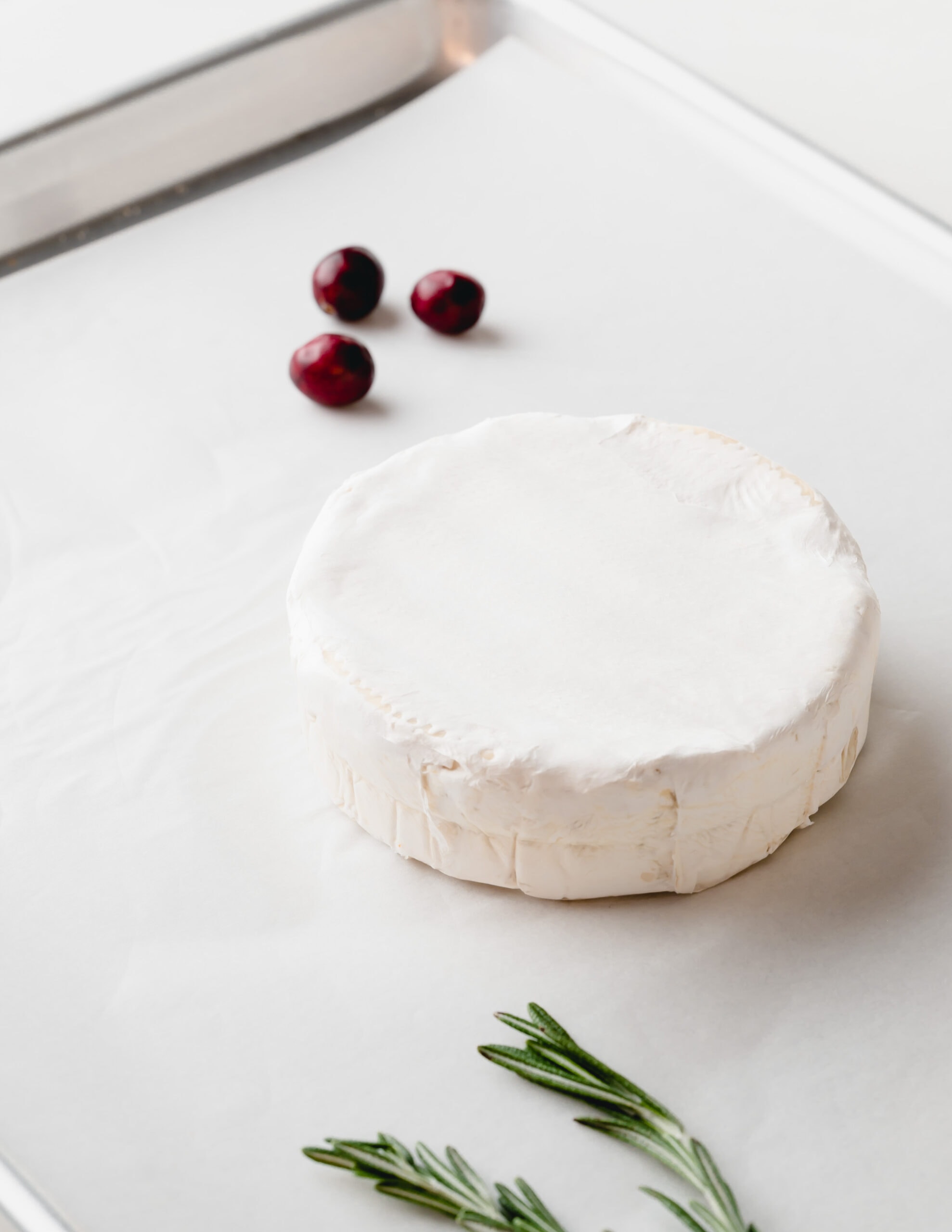 A round of cold brie on a baking sheet with a sprig of rosemary and three cranberries in the back.