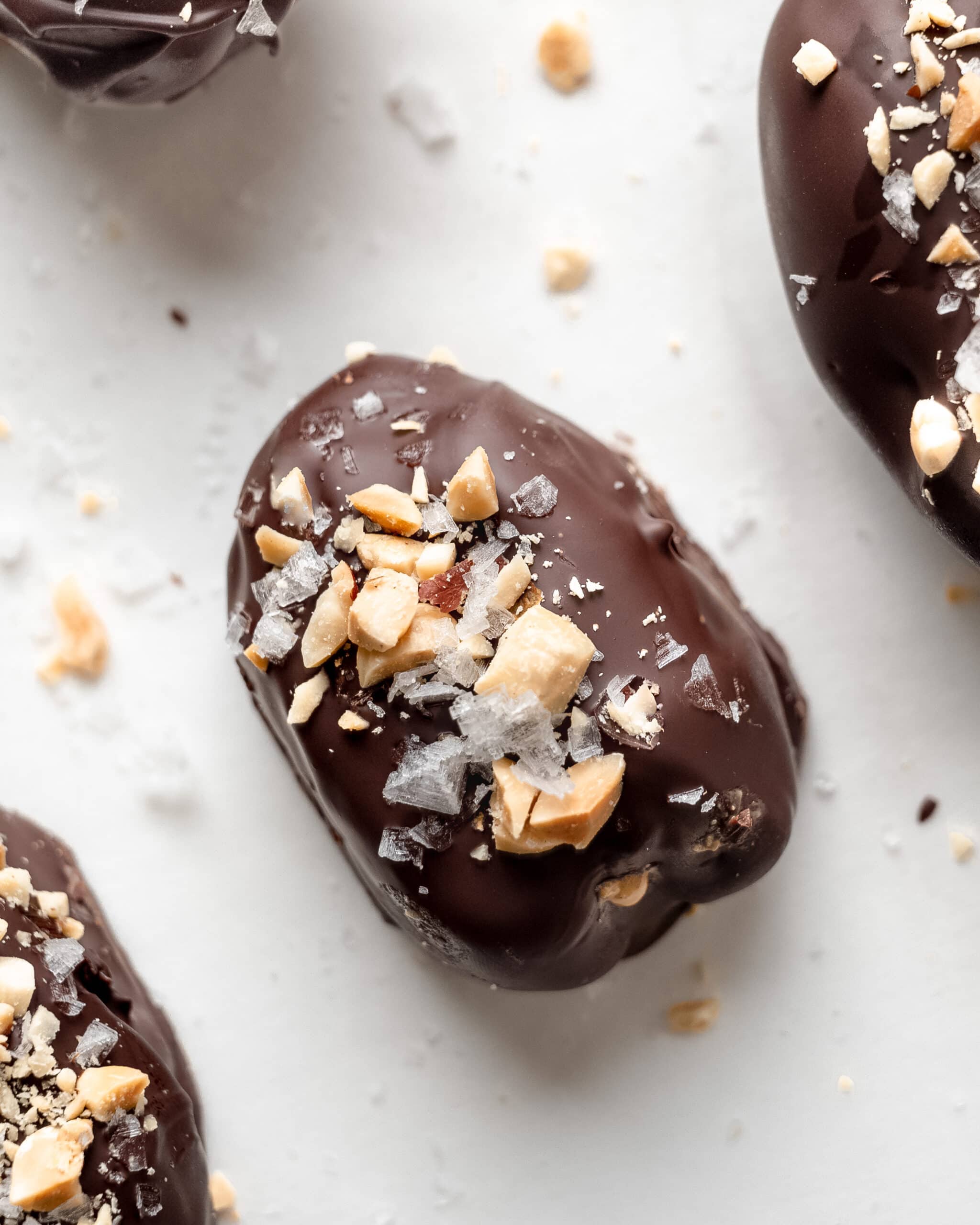 A close up view of a snickers date. Date covered in chocolate with chopped peanuts on top and flaky sea salt. 