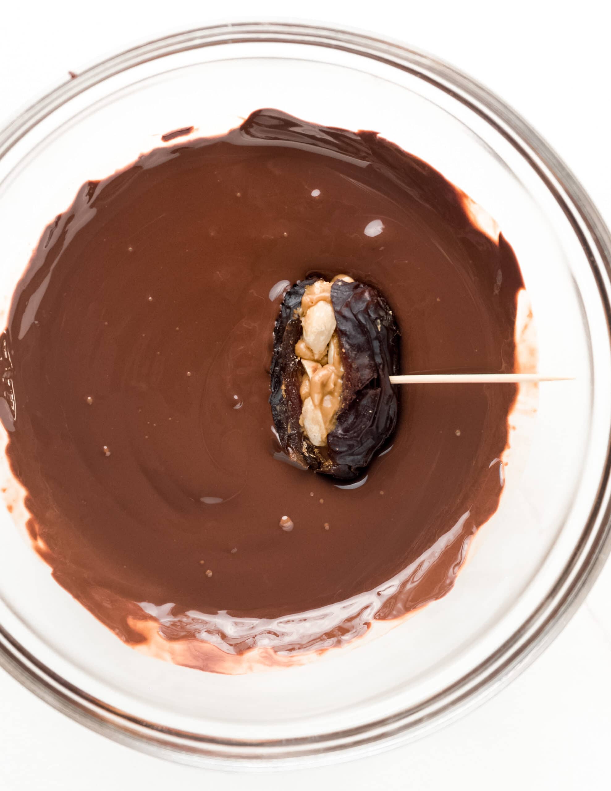 A clear bowl with melted chocolate with a peanut butter stuffed date inside being dipped in the chocolate.