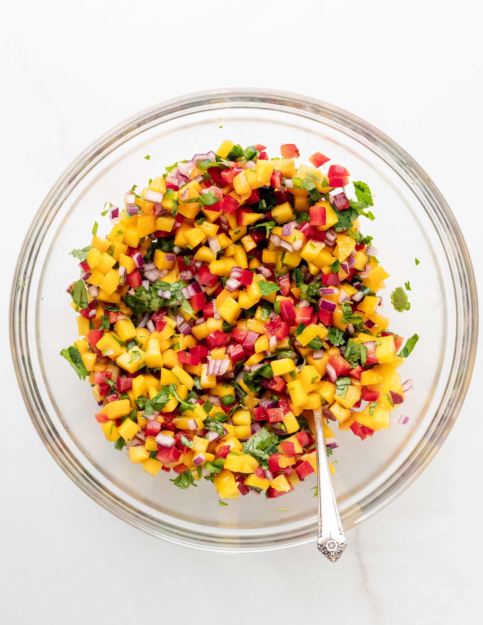 A large clear with with mango salsa ingredients stirred with a silver antique spoon.