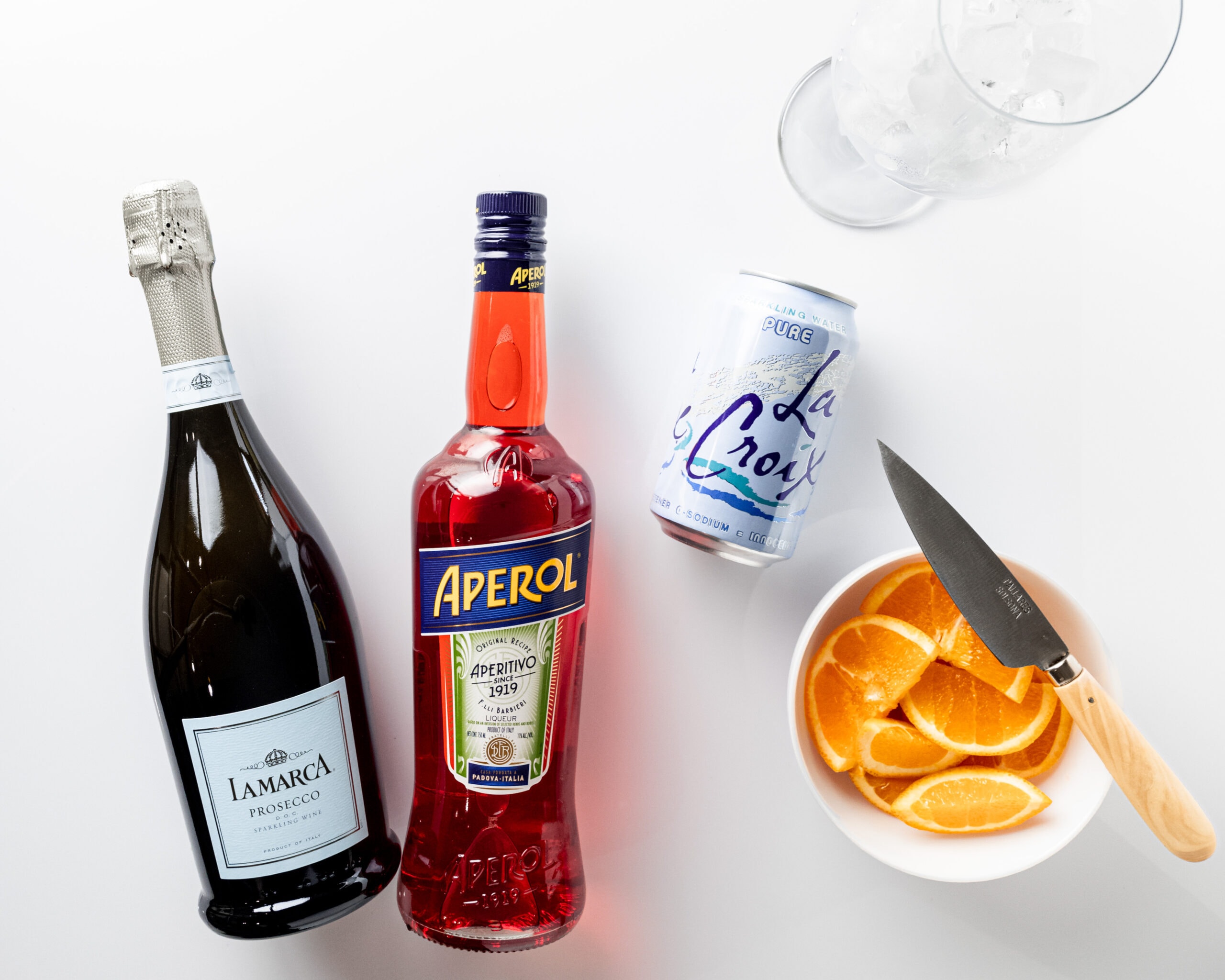 Ingredient shot of aperol spritz. One bottle of Lamarca prosecco, bottle of Aperol, can of sparkling water, a bowl of sliced oranges with a paring knife, and a white wine glass with ice.
