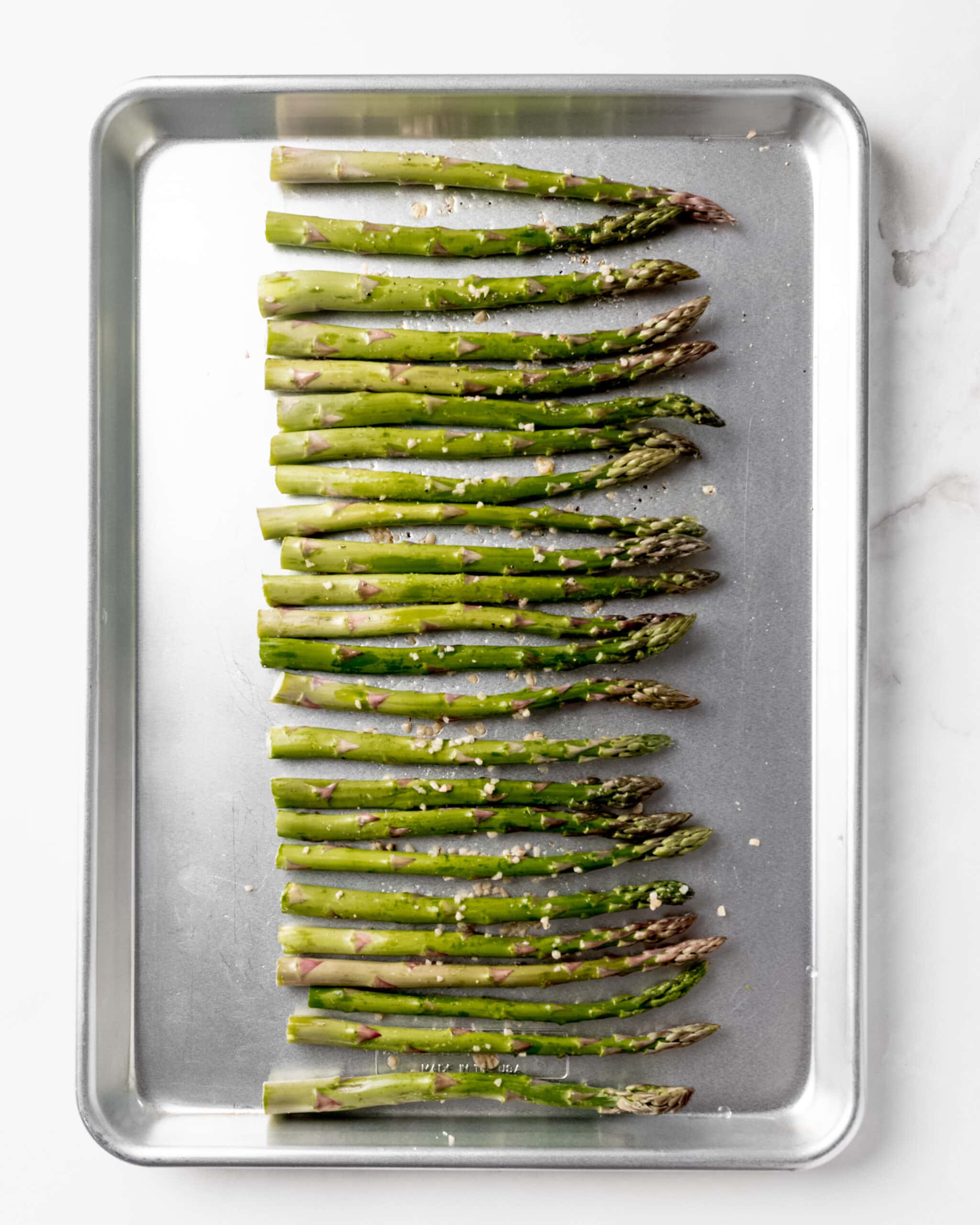 Asparagus on a baking sheet with minced garlic on top, oil, salt, and pepper.