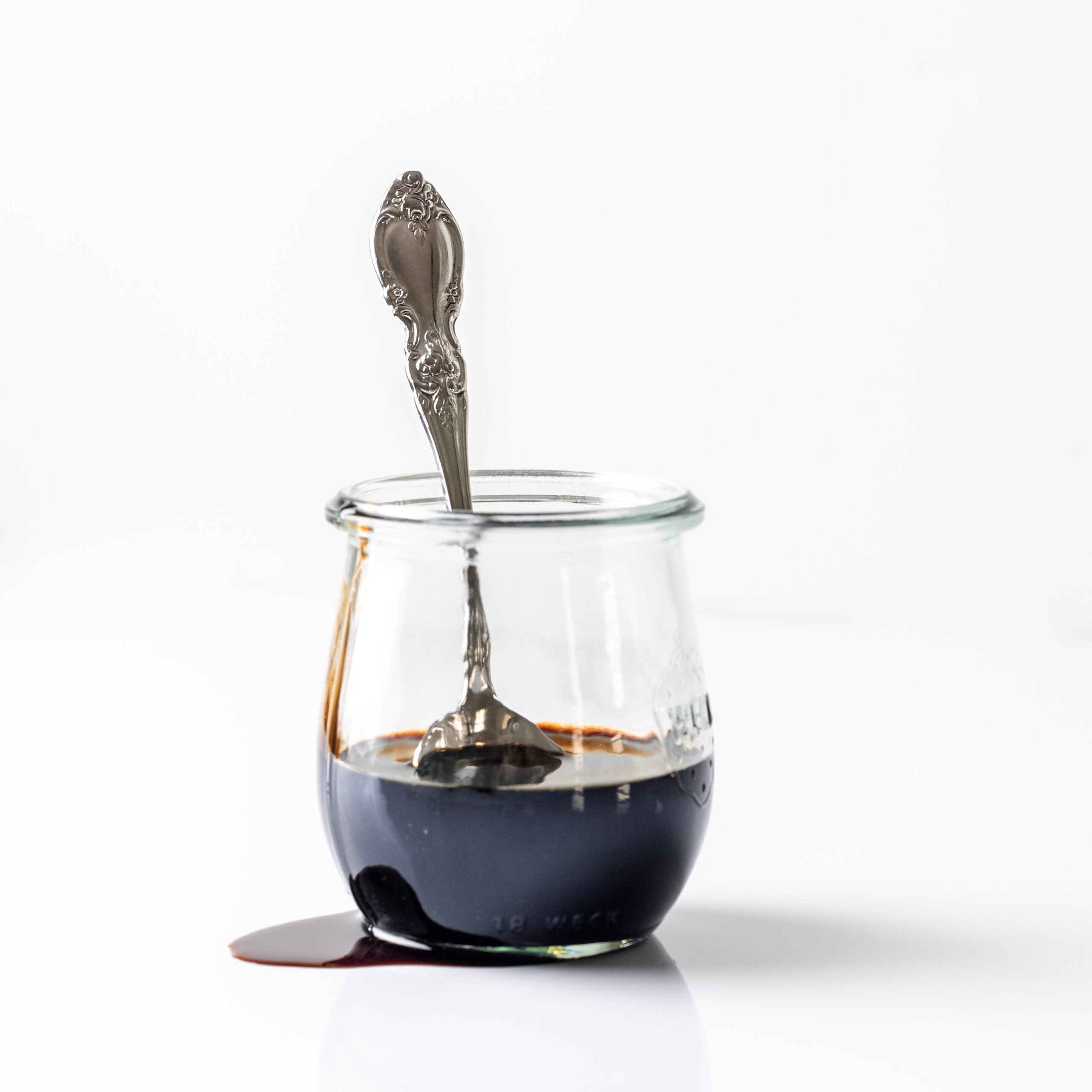 A glass jar with balsamic glaze dripping off the side of the jar with a large silver spoon inside the jar.