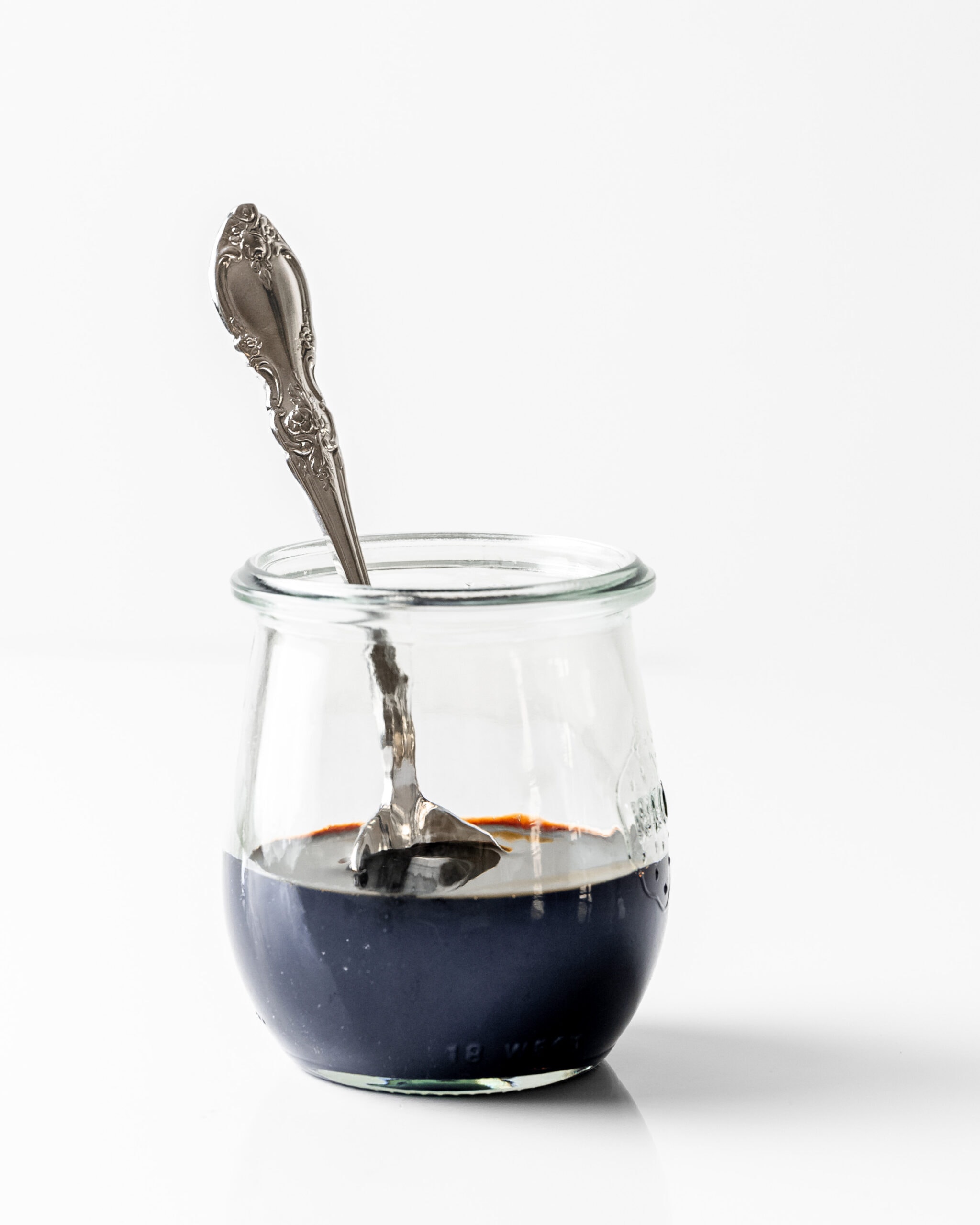 A picture of a glass jar of balsamic glaze with a silver spoon inside the jar.