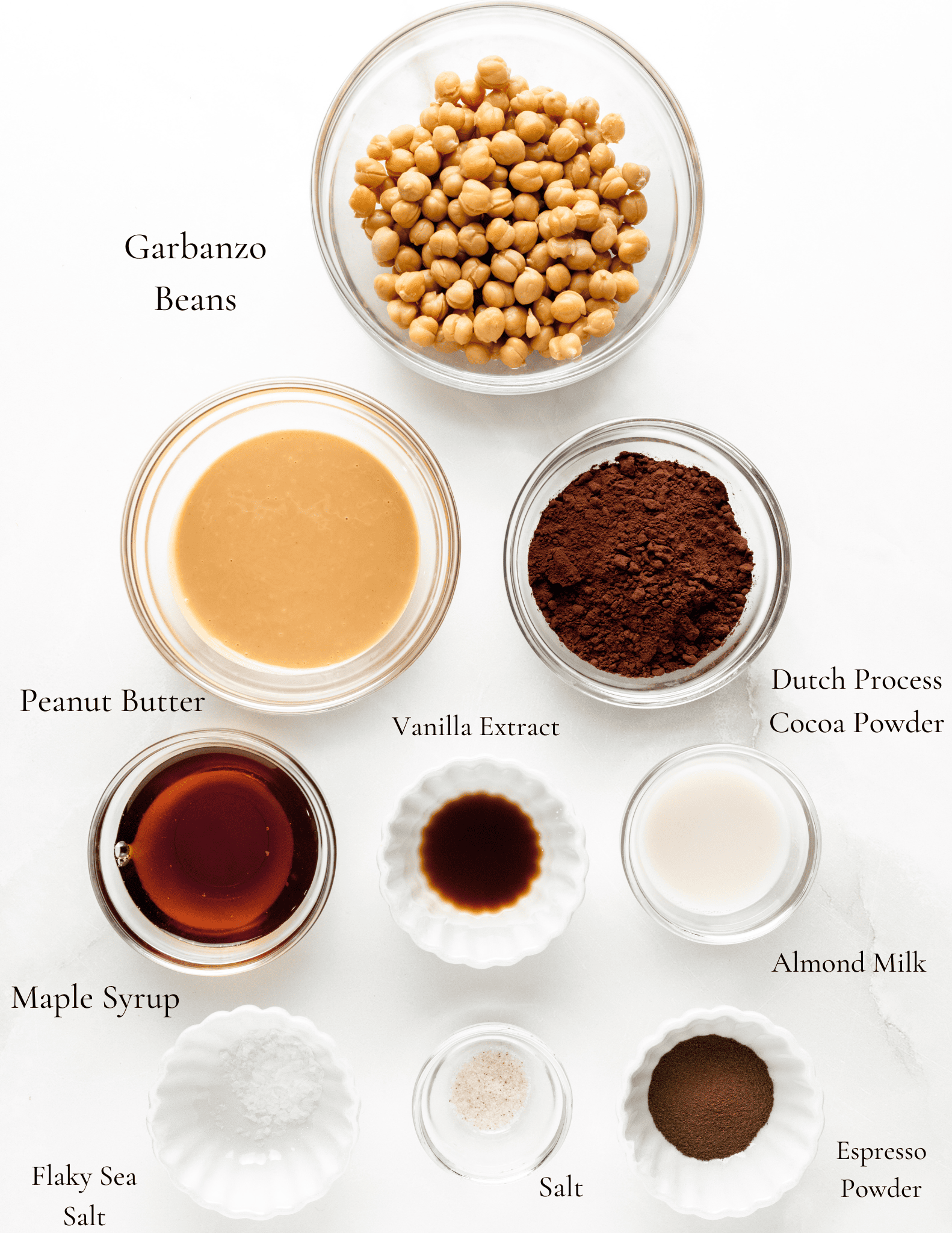 Picture of dark chocolate hummus ingredients, including a bowl of garbanzo beans, dark dutch process cocoa powder, peanut butter, vanilla extract, maple syrup, almond milk, espresso powder, salt, and flaky sea salt. 