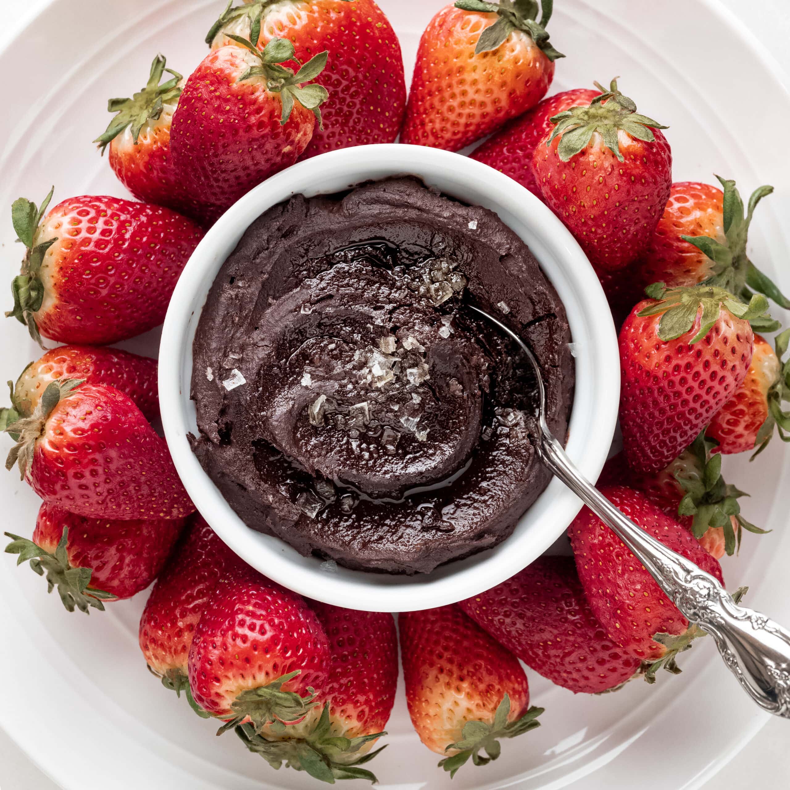 Dark chocolate hummus in a white bowl with sea salt on top, and a silver spoon on the side with red strawberries surrounding the bowl.
