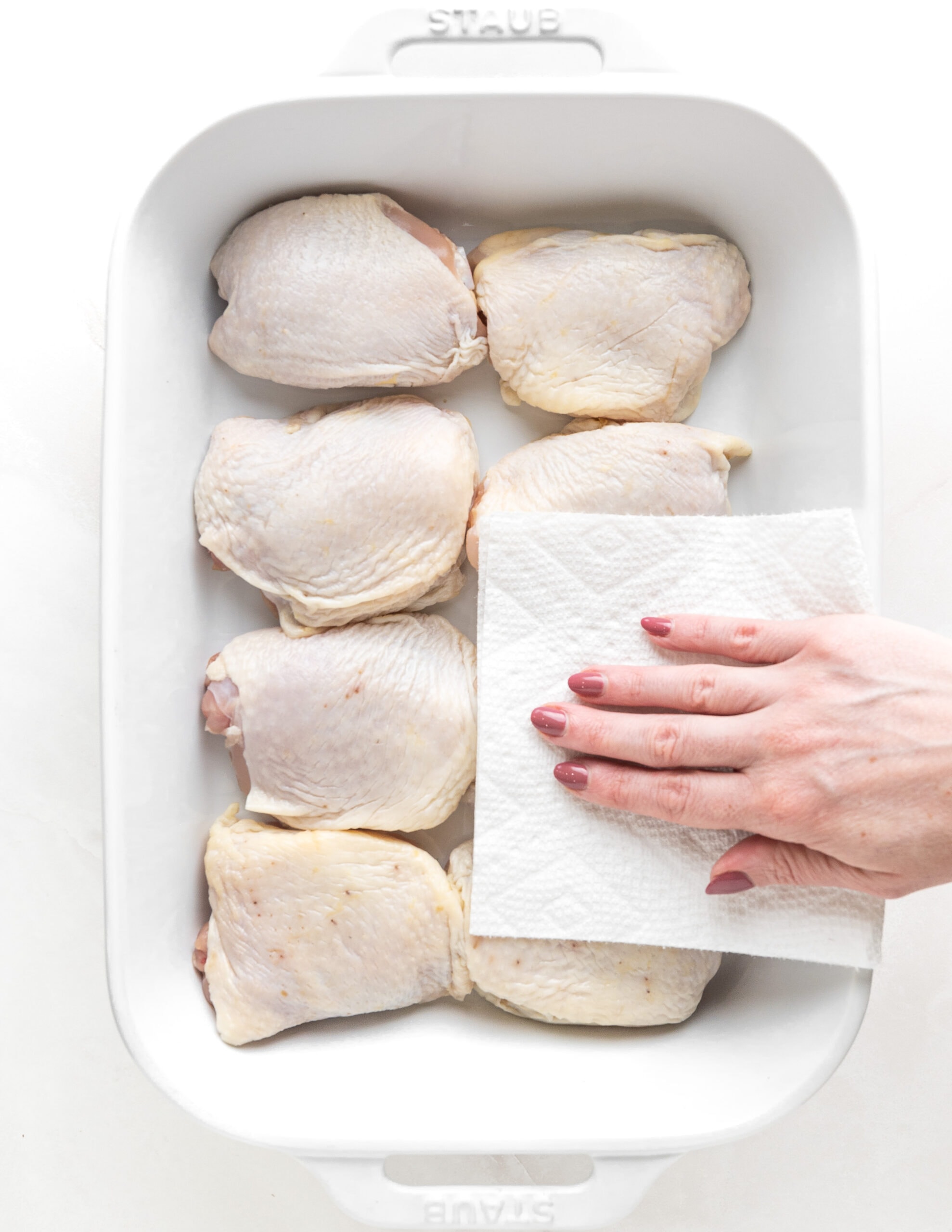 A large white ceramic rectangular baking dish with 8 bone-in chicken thighs, and a hand off to the side drying the chicken with a paper towel. 