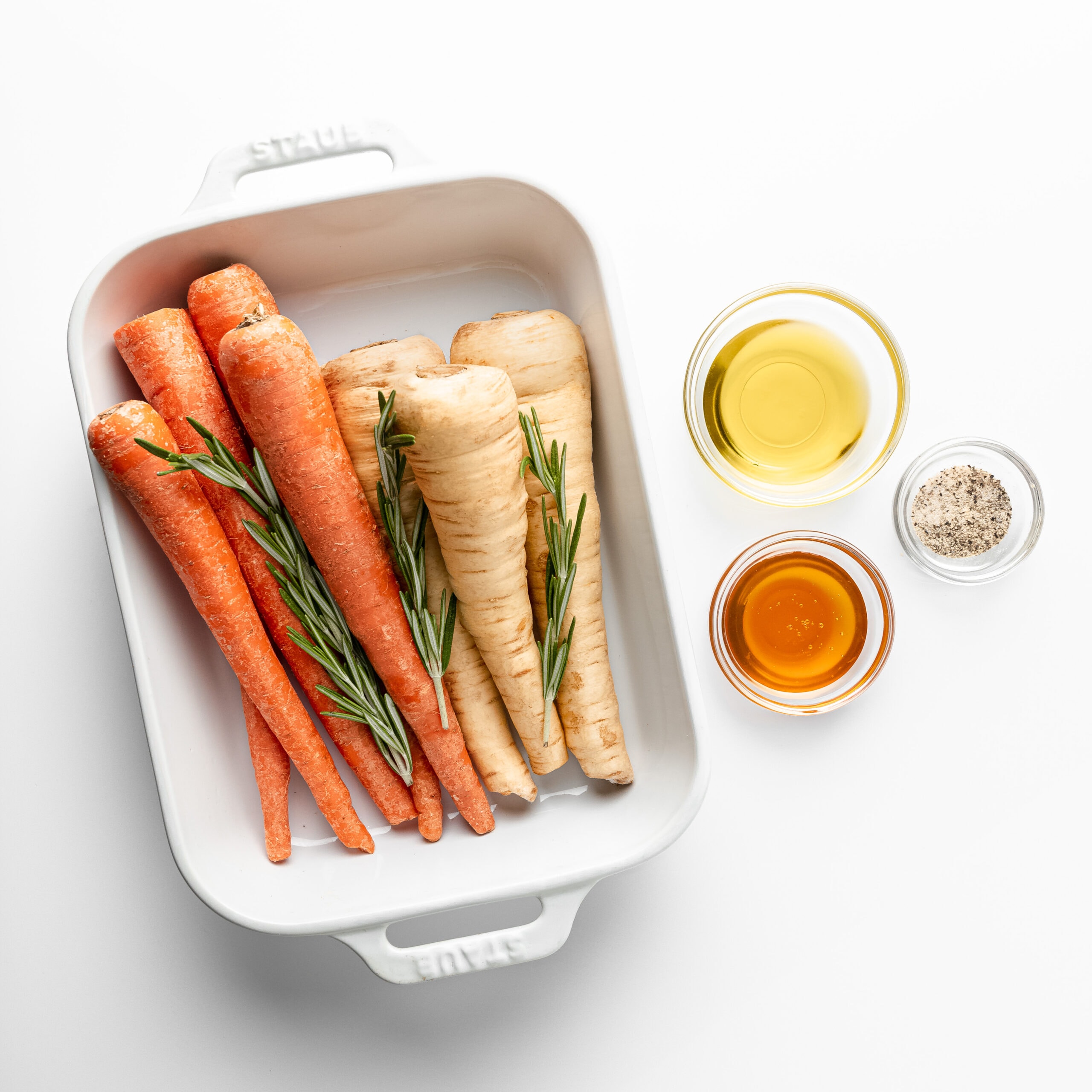A white ceramic dish with carrots, parsnips, and fresh rosemary. Off to the side of the dermaic dish are three glass bowls filled with olive oil, spices, and honey.