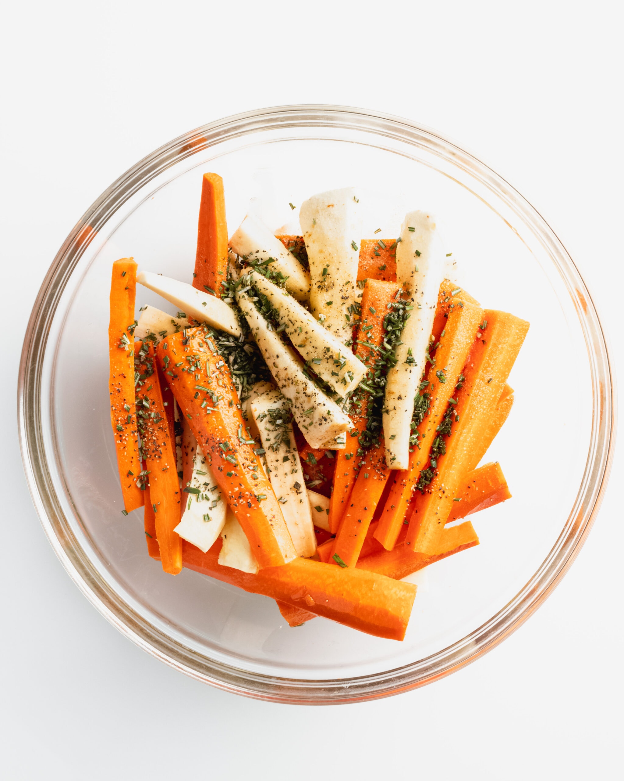 A clear glass bowl with chopped and peeled carrots, parsnips, and fresh herbs. 