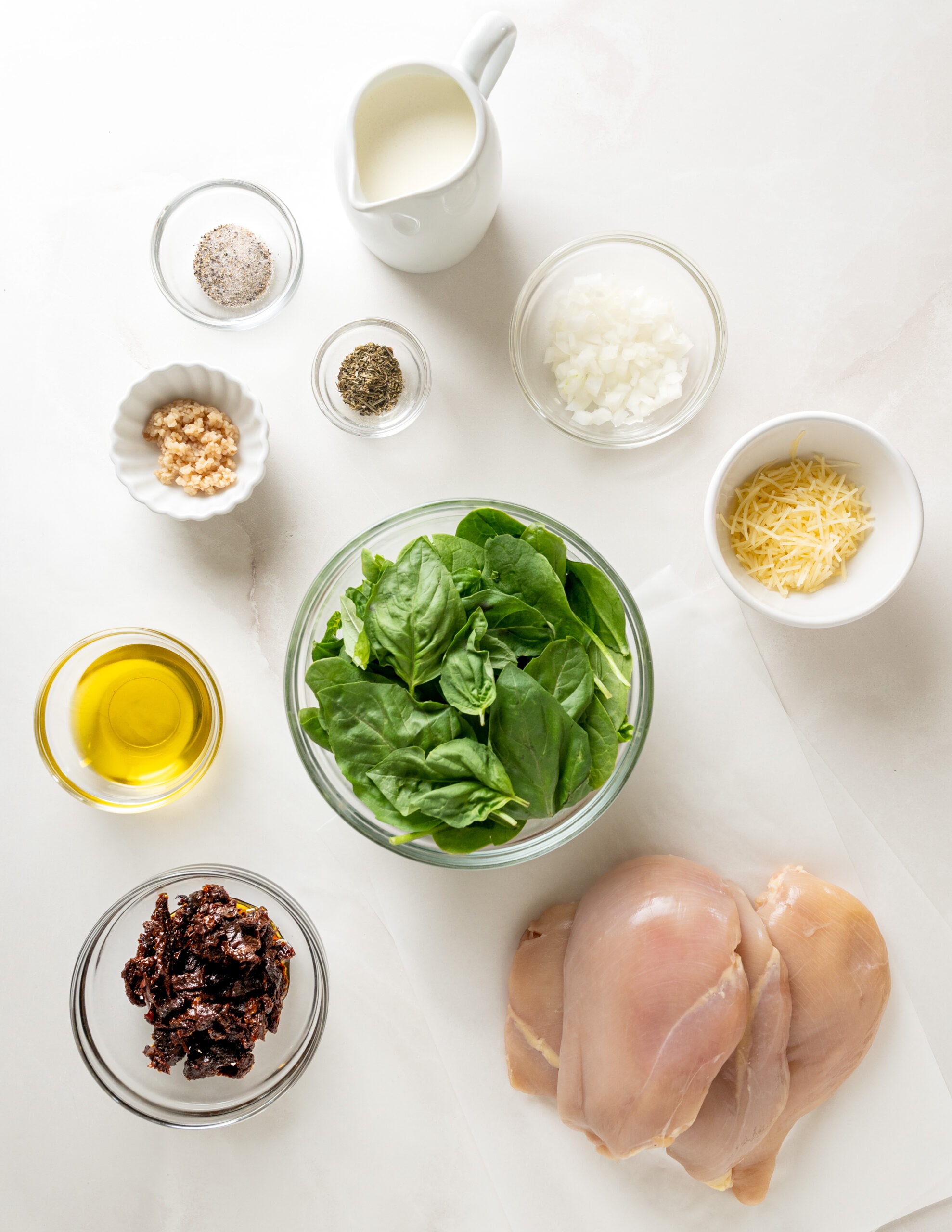 Ingredients for tuscan chicken. A large clear bowl of spinach and basil, alongside a bowl of sundried tomatoes, olive oil, parmesan cheese, yellow onion, heavy cream, minced garlic, salt, pepper, italian seasoning, and chicken breasts. 