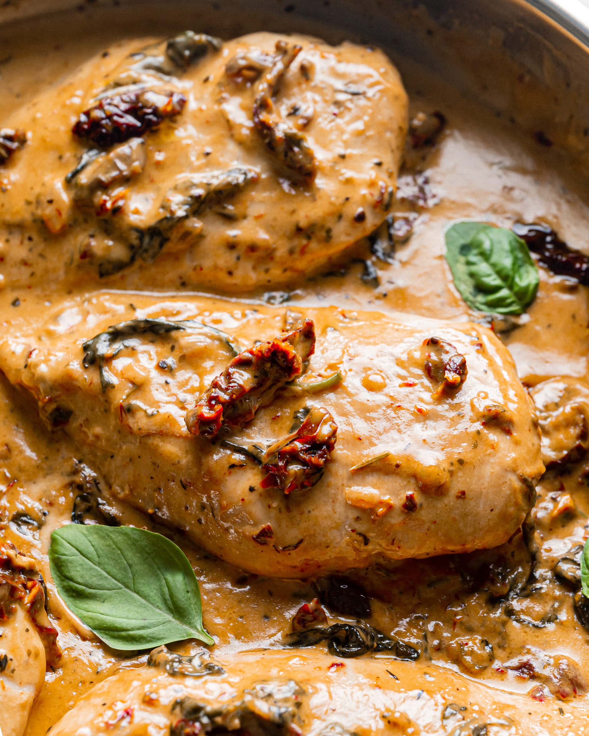 Three pieces of tuscan chicken, coated in a rich tomato cream sauce with fresh basil leaves and sundried tomatoes on top.