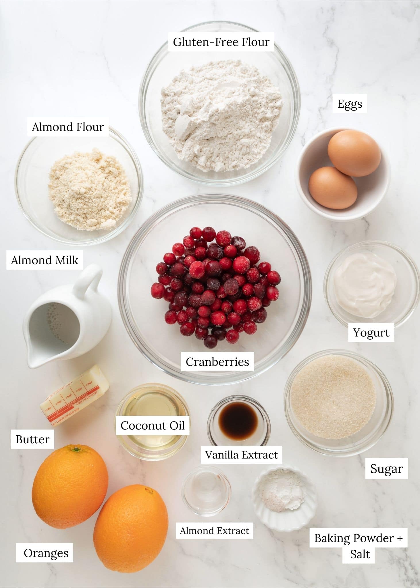 Cranberry orange muffin ingredients. A large clear bowl with cranberries next to a bowl with flour, almond flour, eggs, yogurt, milk, butter, coconut oil, navel oranges, extracts, baking powder, salt, and sugar.