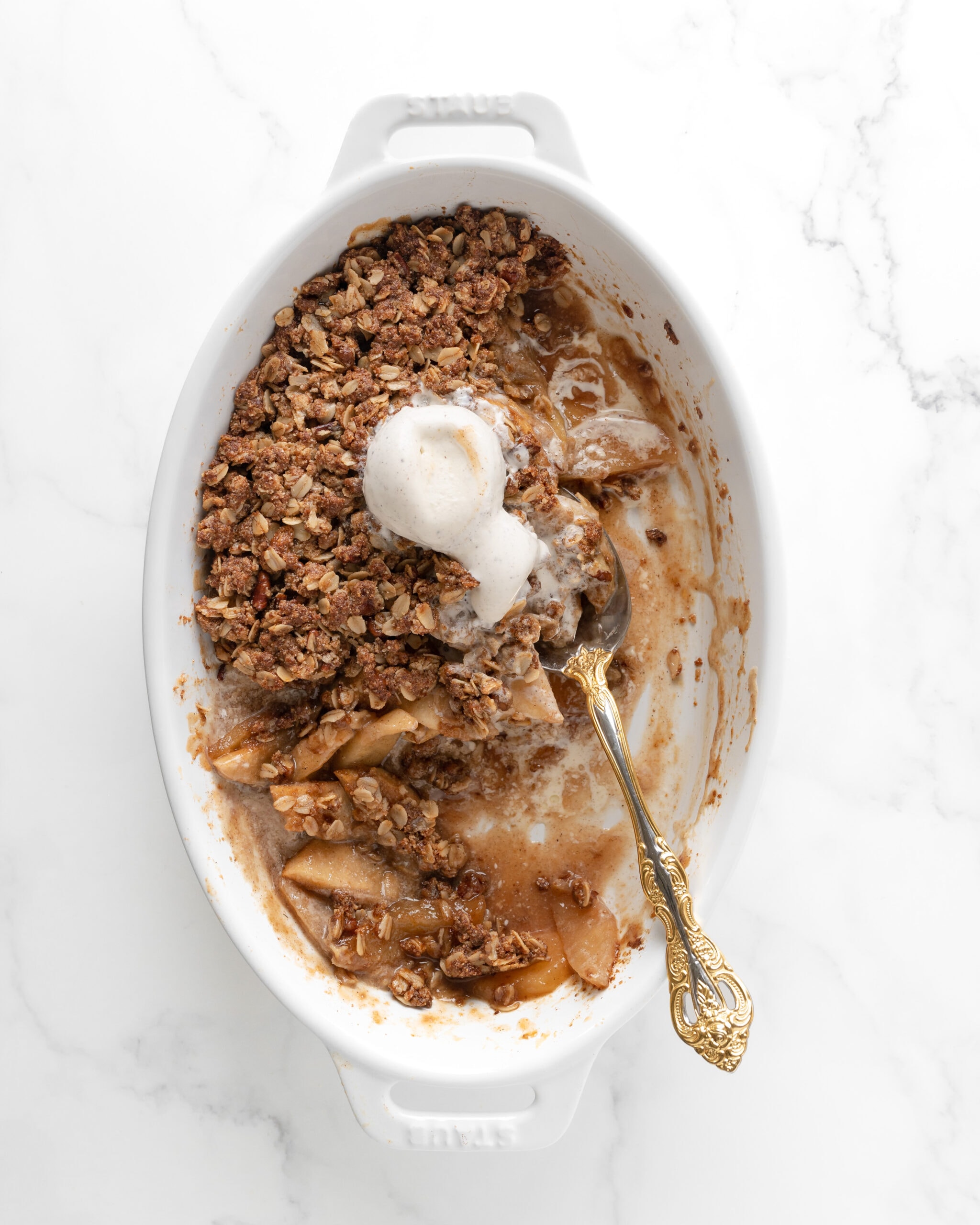 An oval baker with healthy apple crisp melted and half eaten with a spoon in the middle of the crisp.