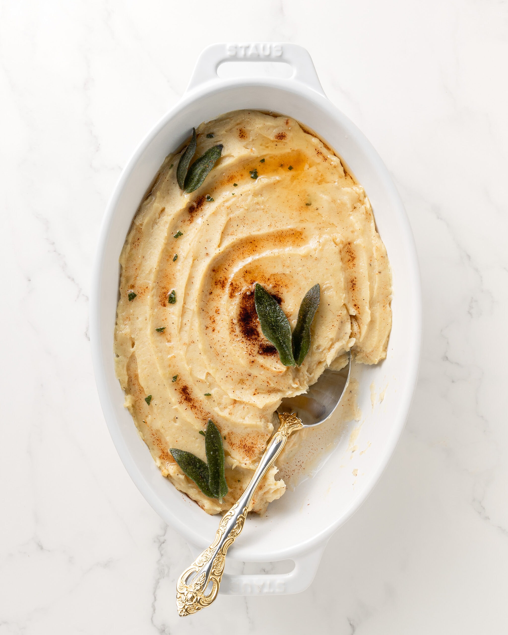 White mashed sweet potatoes in an oval white dish with sage leaves and a gold and silver spoon scooping out some of the potatoes. 