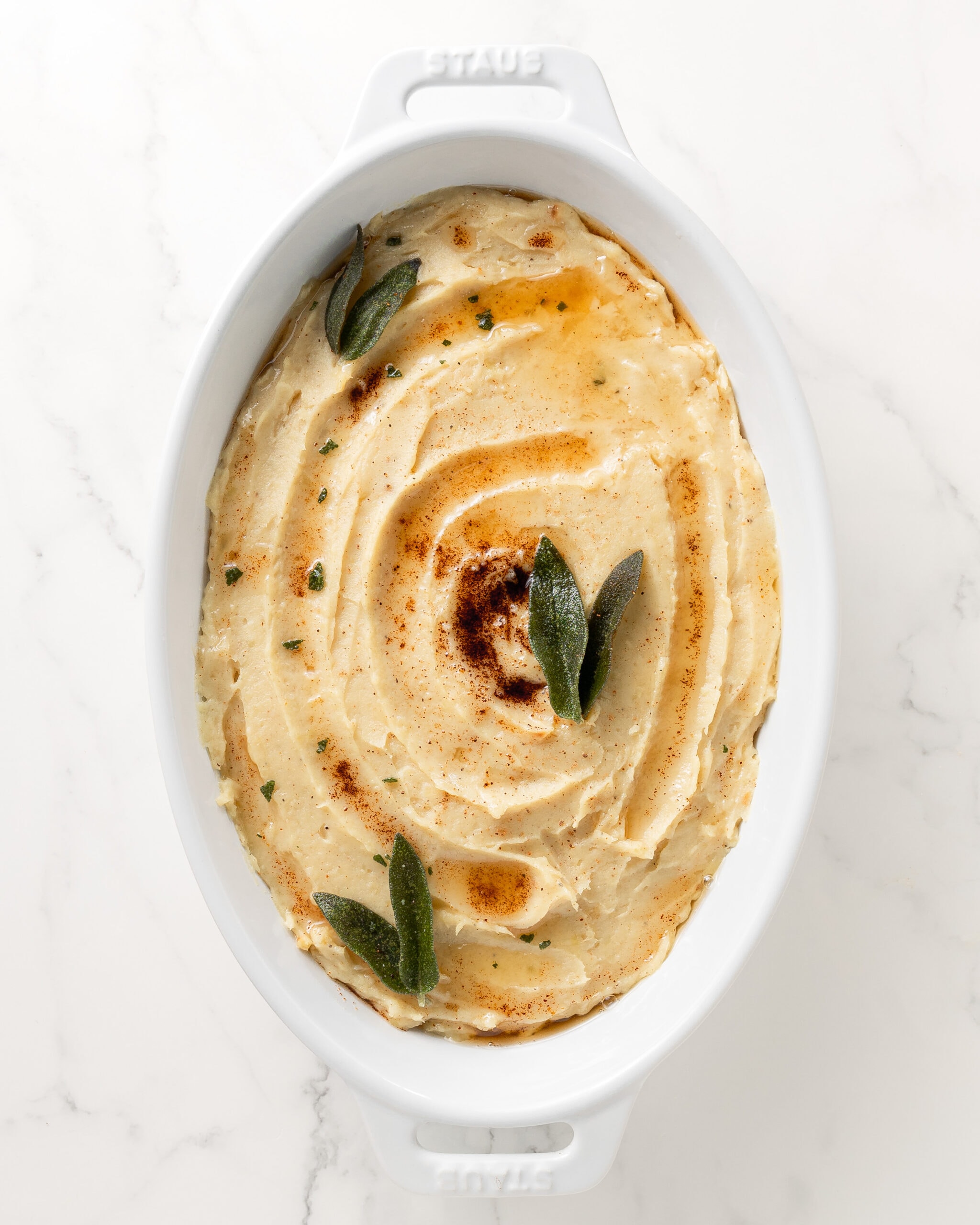 Mashed white sweet potatoes in an oval white bakers dish with brown butter and sage leaves on top.