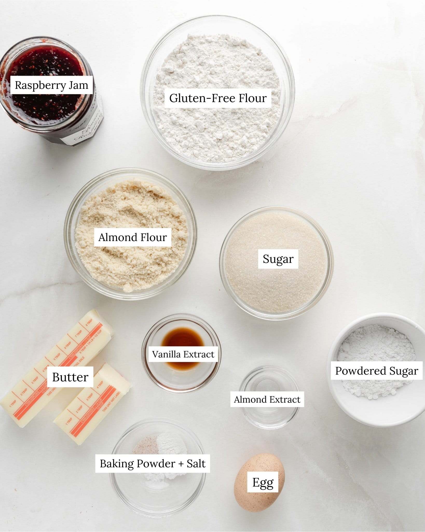Ingredients for gluten-free linzer cookies. Gluten-free flour in a clear bowl, sugar, vanilla extract, almond extract, baking powder, salt, butter, egg, almond flour, and powdered sugar.
