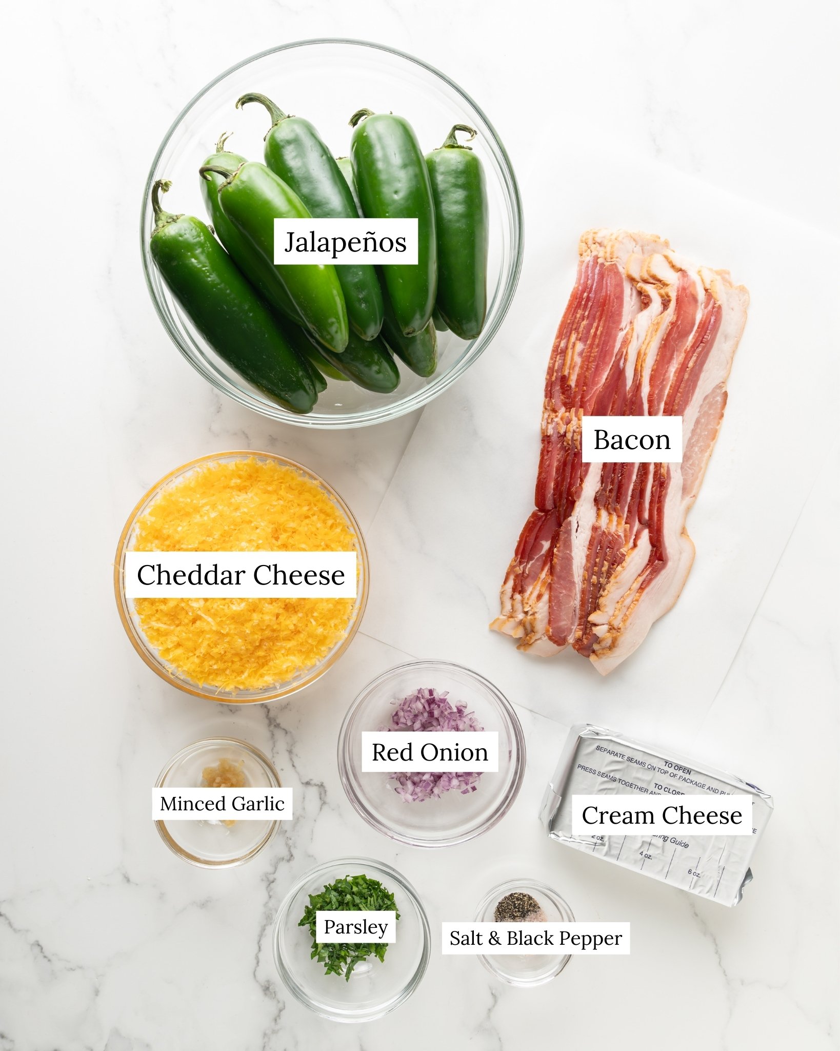 Low carb jalapeno popper ingredients. A medium sized clear bowl of fresh jalapenos, slices of bacon, grated cheese, red onion, minced garlic, minced parsley, cream cheese, salt and pepper.