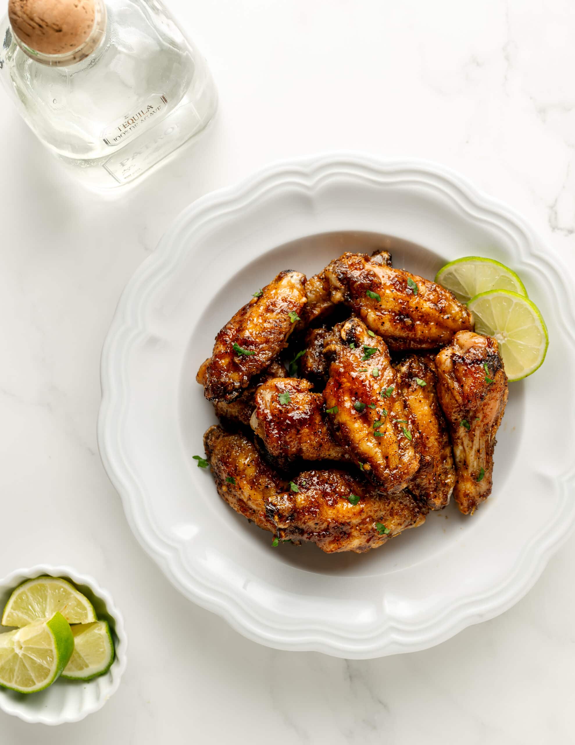 Tequila Lime Chicken Wings in a white scalloped bowl with two lime wedges, garnishes with a sprinkle of cilantro. In the corner is a bottle of Patron and opposite, a bowl of lime wedges. 