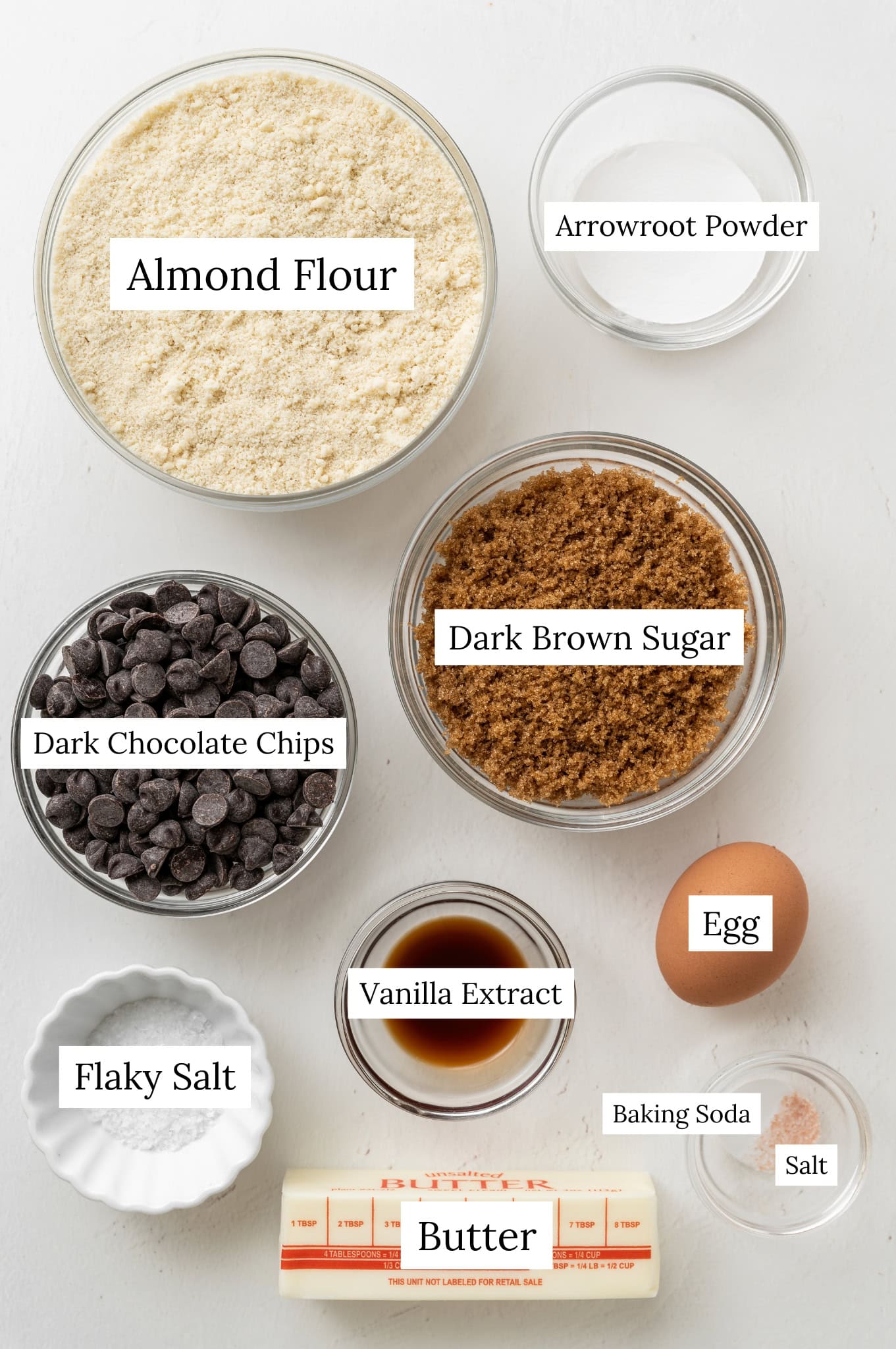 Ingredients to make almond flour chocolate chip cookies. A bowl of almond flour, arrowroot powder, dark brown sugar, dark chocolate chips, egg, vanilla extract, baking soda, salt, butter, and flaky salt.