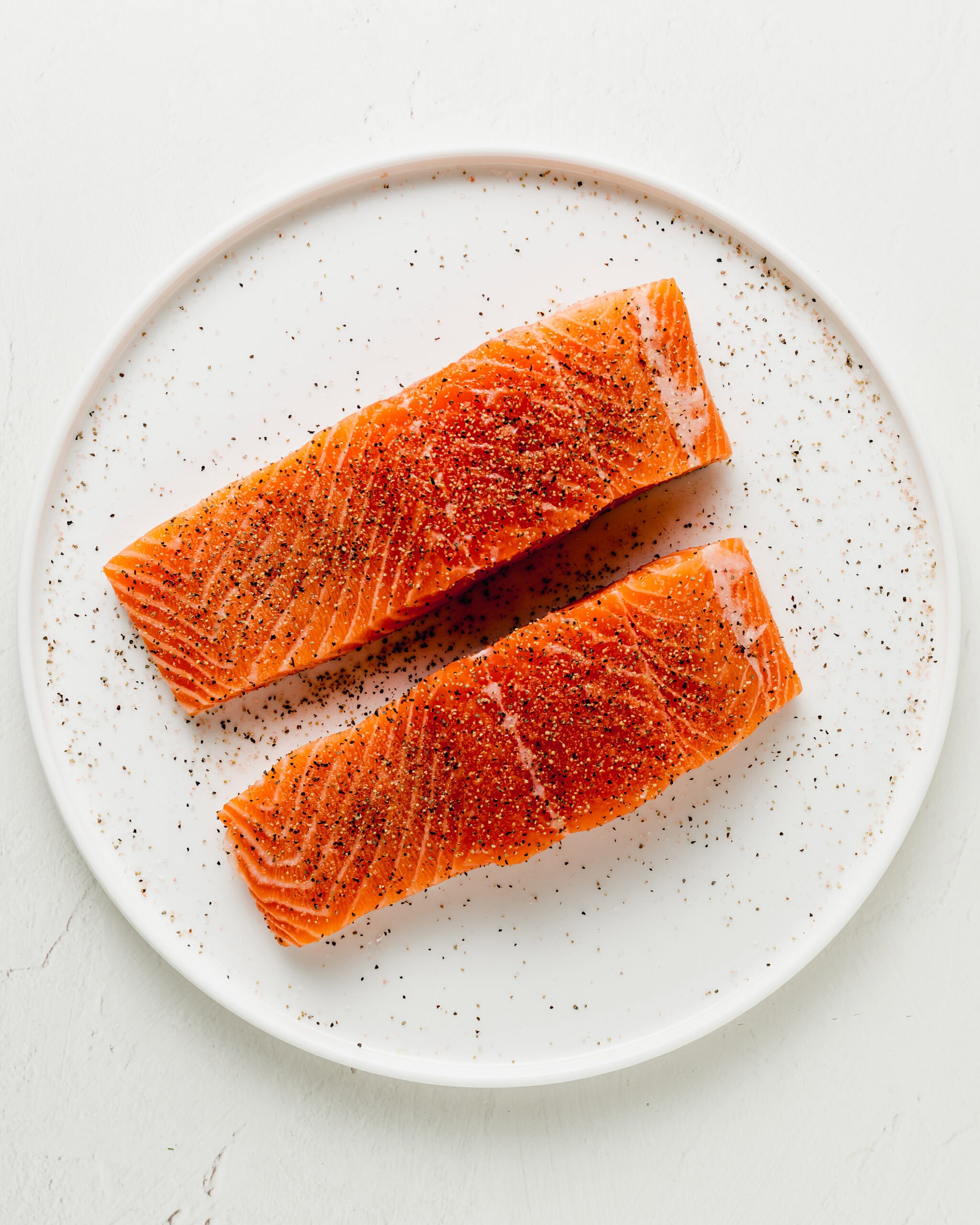 Salmon on a white plate seasoned generously with salt and black pepper.