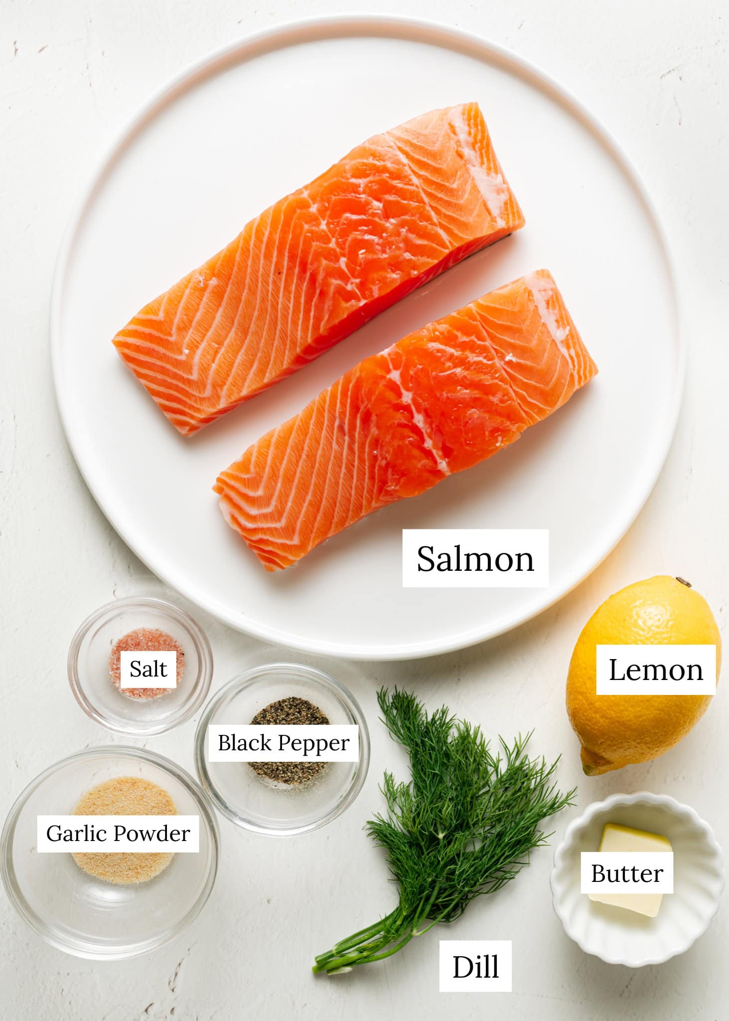Ingredient shot of baked salmon. Salmon on a white plate, lemon, salt, black pepper, garlic powder, butter, and dill.
