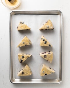 Blueberry scones on a lined baking sheet, applying an egg wash on top.