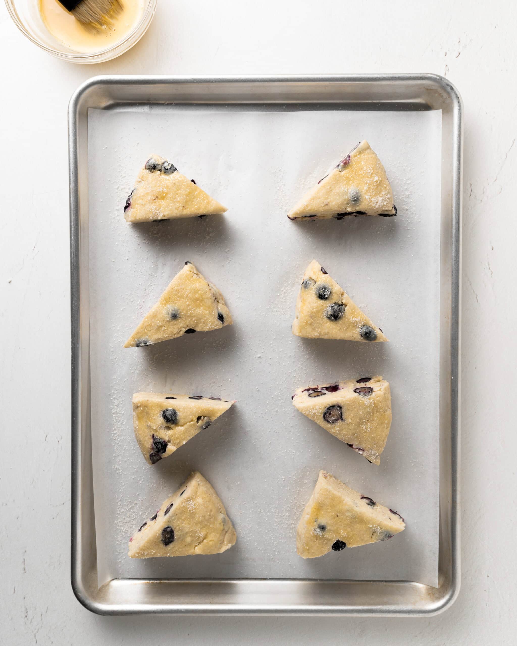 Blueberry scones on a lined baking sheet, applying an egg wash on top.