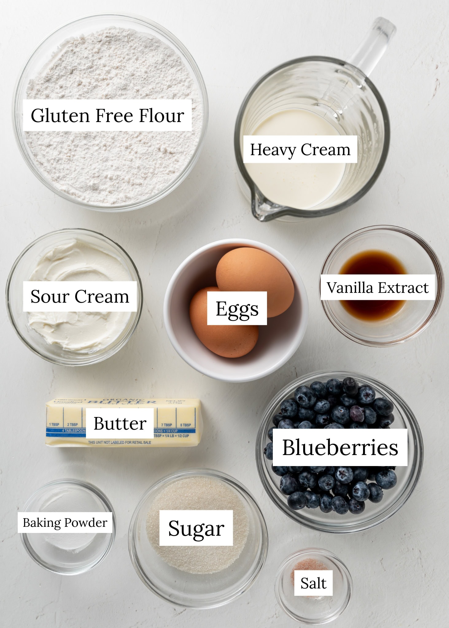 Blueberry scone ingredients. A bowl of gluten free flour, cup of heavy cream, sour cream, eggs, vanilla extract, butter, baking powder, sugar, blueberries, salt.