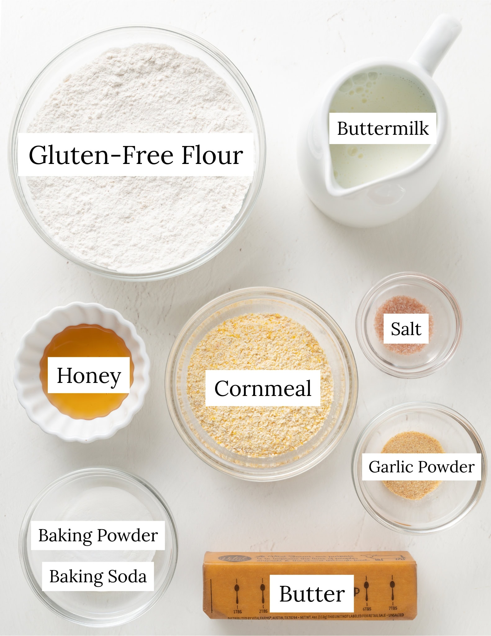 Ingredients for cornmeal biscuits. Buttermilk, gluten-free flour, cornmeal, salt, baking powder, baking soda, garlic powder, honey, and butter.
