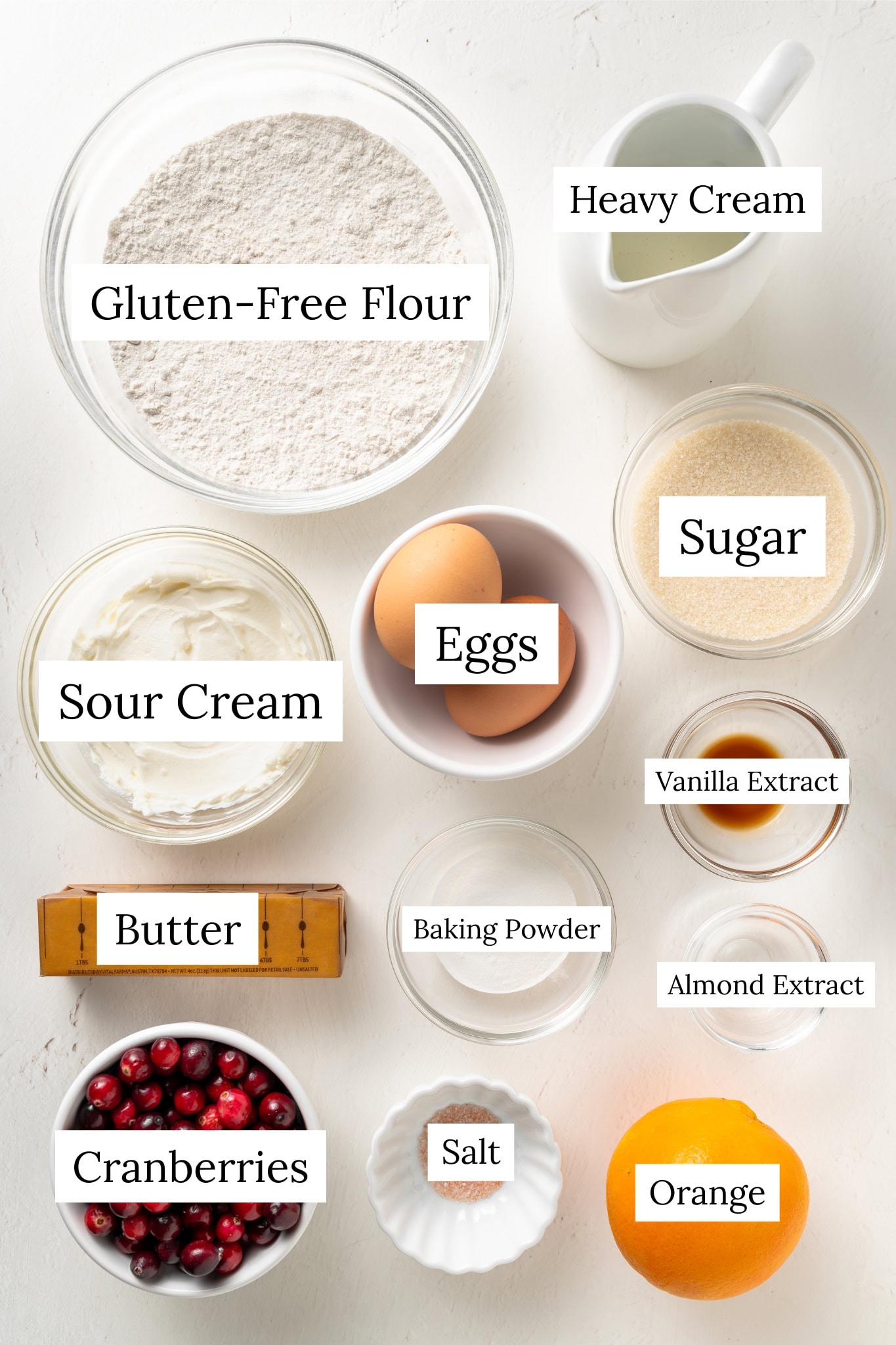 Overhead shot of all the ingredients for the Cranberry Orange Scones recipe, including a large bowl of gluten-free flour, sour cream, two eggs, granulated sugar, whole cranberries, a whole orange, heavy cream in a small pitcher, and small bowls containing vanilla extract, almond extract, and baking powder/salt.