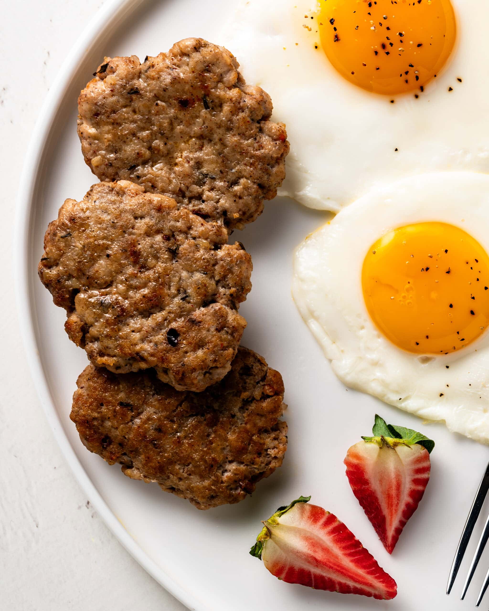 Large white plate with 3 chicken breakfast sausage patties, two large eggs with black pepper, and a strawberry cut in half.
