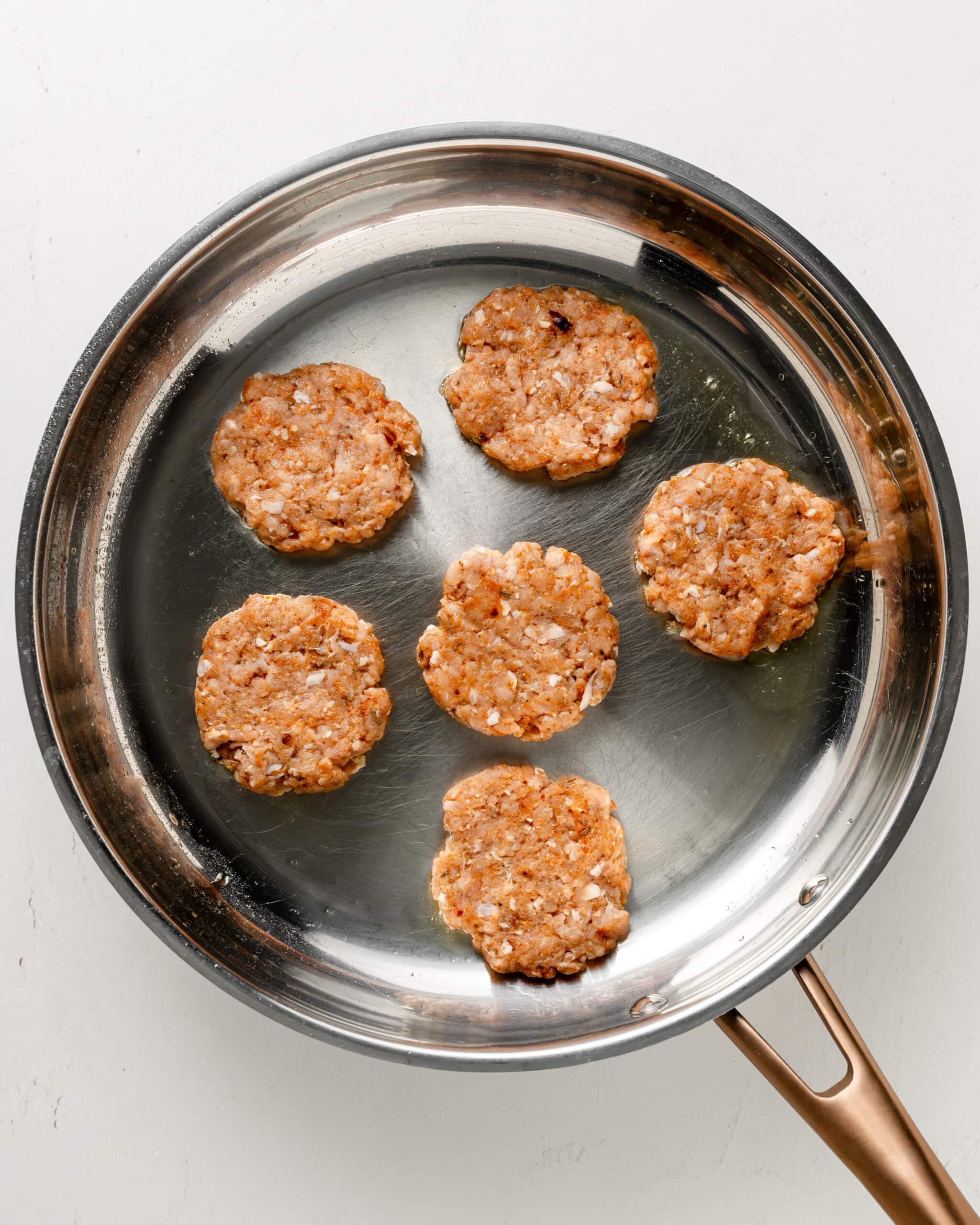 Ground chicken sausage patties frying in a large stainless steel pan.