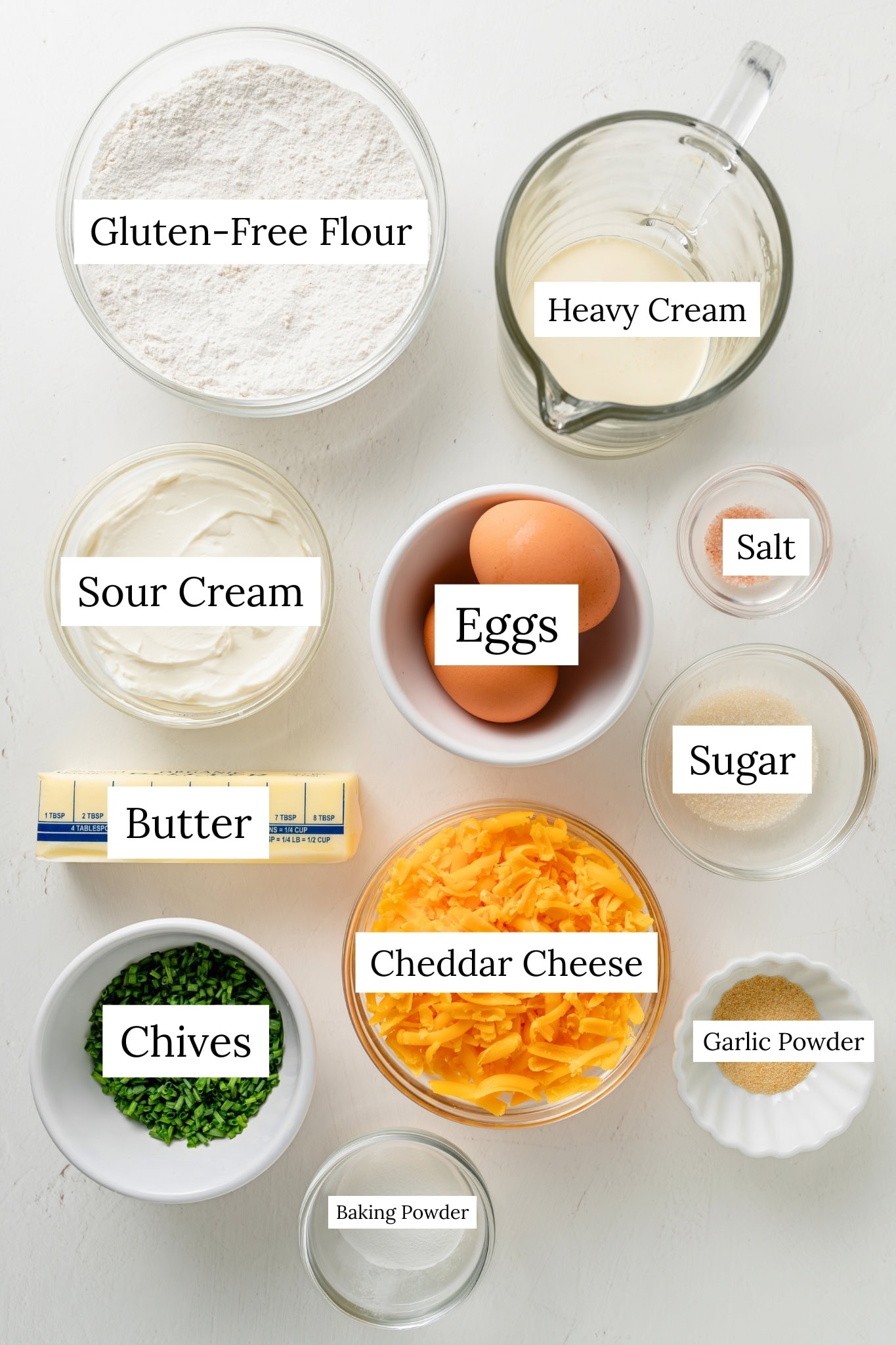 Ingredients for cheddar scones. Gluten free flour, heavy cream, sour cream, eggs, salt, sugar, cheddar cheese, chives, butter, garlic powder, baking powder.
