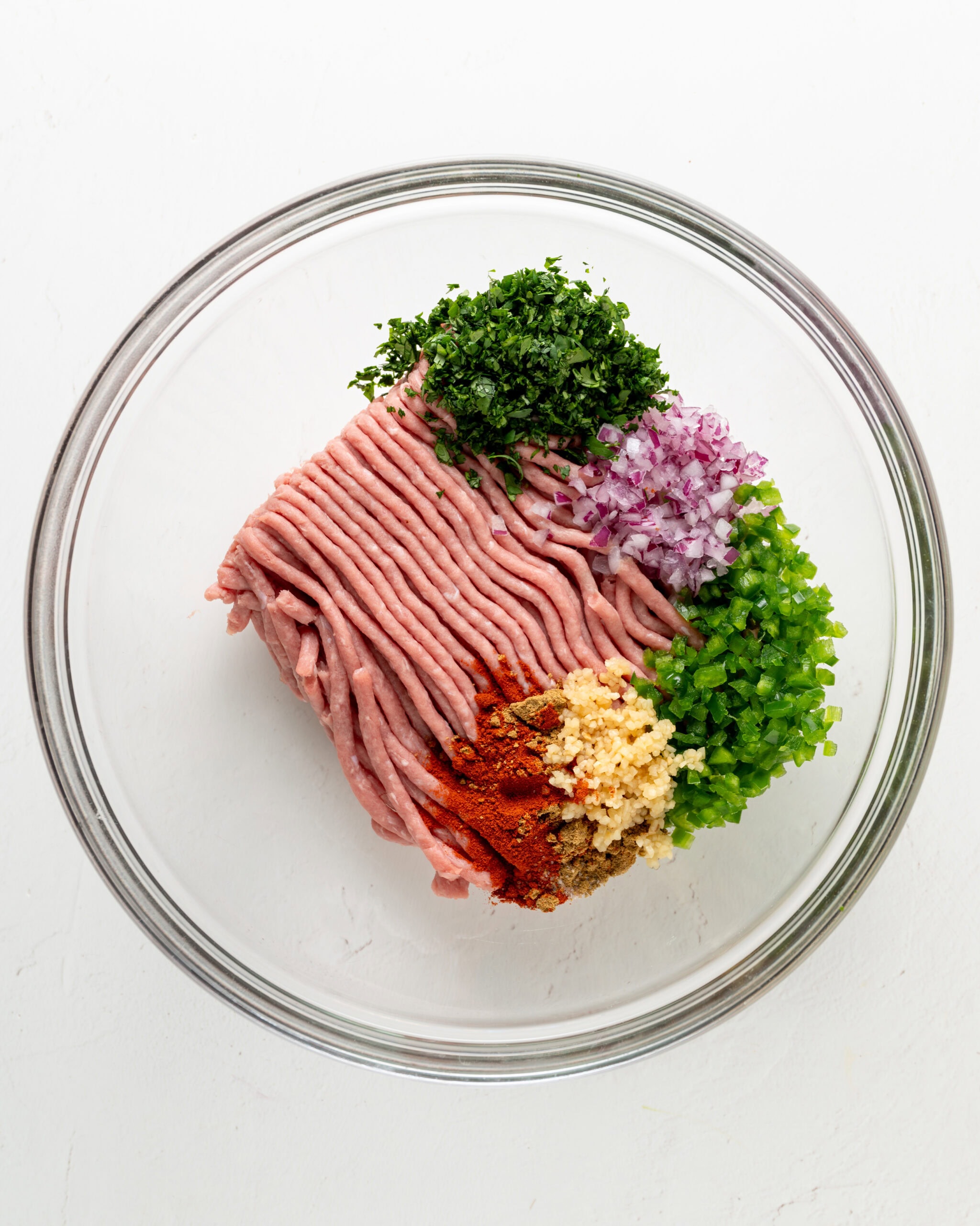 A glass mixing bowl containing ground turkey, diced red onions, fresh cilantro, minced garlic, and sliced jalapeños ready to be mixed.
