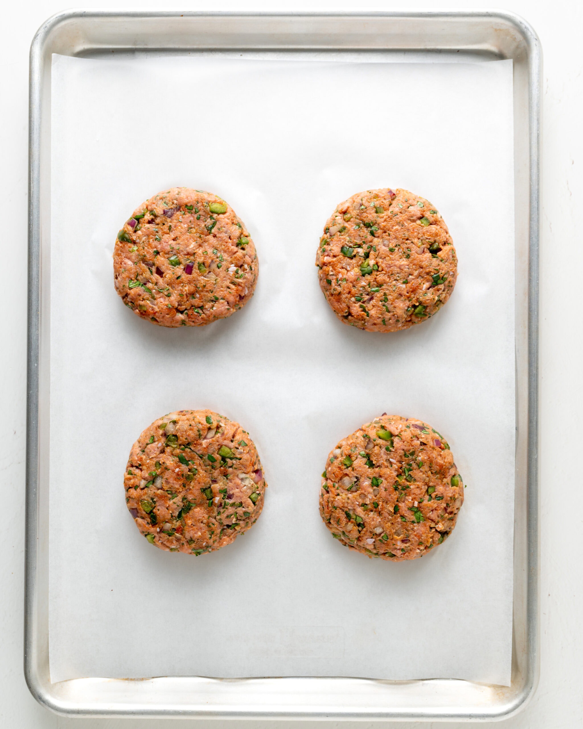 Four even turkey burger patties arranged on a parchment-lined baking sheet, showing flecks of fresh herbs and spices.