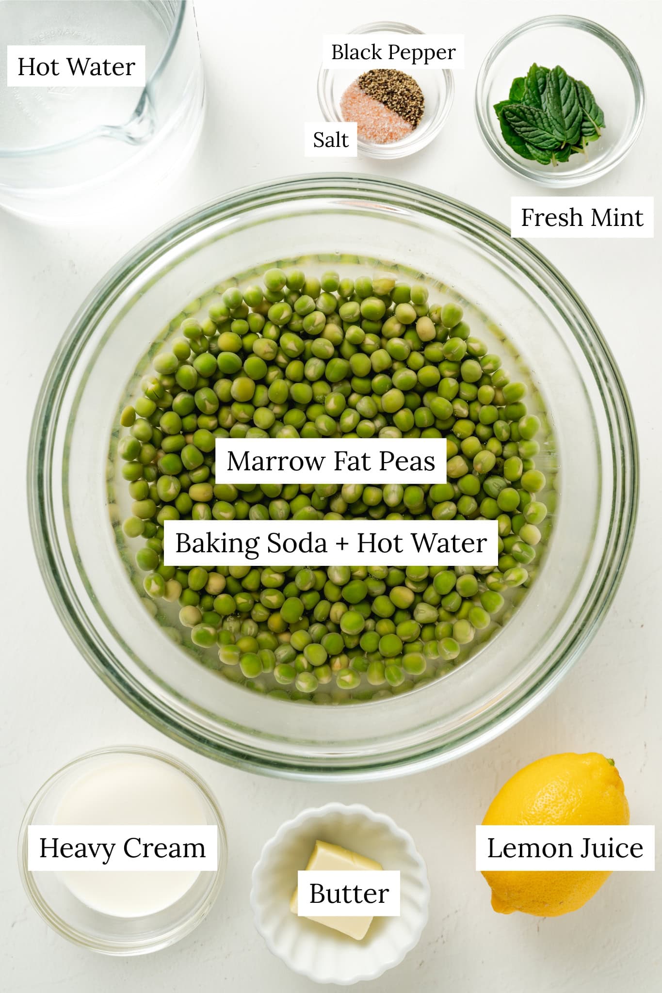 In the center is a large glass bowl containing green marrowfat peas soaking in water. Surrounding the bowl are several small prep dishes: one with fresh mint leaves, another with a blend of pink salt and black pepper, a small bowl of heavy cream, and a fluted dish containing a square pat of butter. A whole yellow lemon and a large glass pitcher of water are also positioned around the main bowl, creating a bright and organized culinary display.