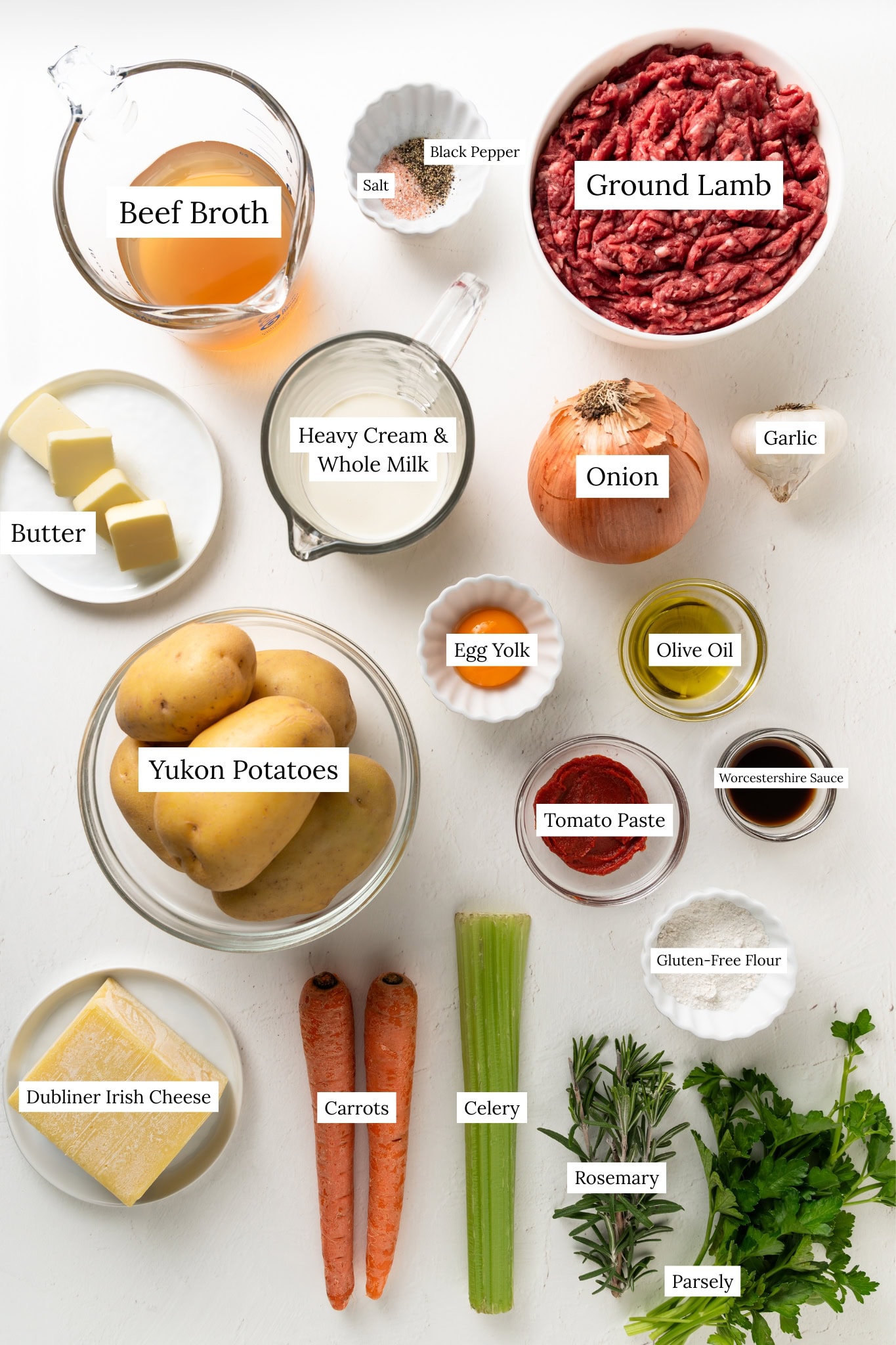 A flat-lay ingredient shot for gluten-free Shepherd’s Pie on a white background. Ingredients include a bowl of ground lamb, Yukon Gold potatoes, carrots, celery, onion, garlic, Dubliner Irish cheese, beef broth, milk, butter, an egg yolk, tomato paste, olive oil, gluten-free flour, Worcestershire sauce, and fresh herbs like rosemary and parsley.