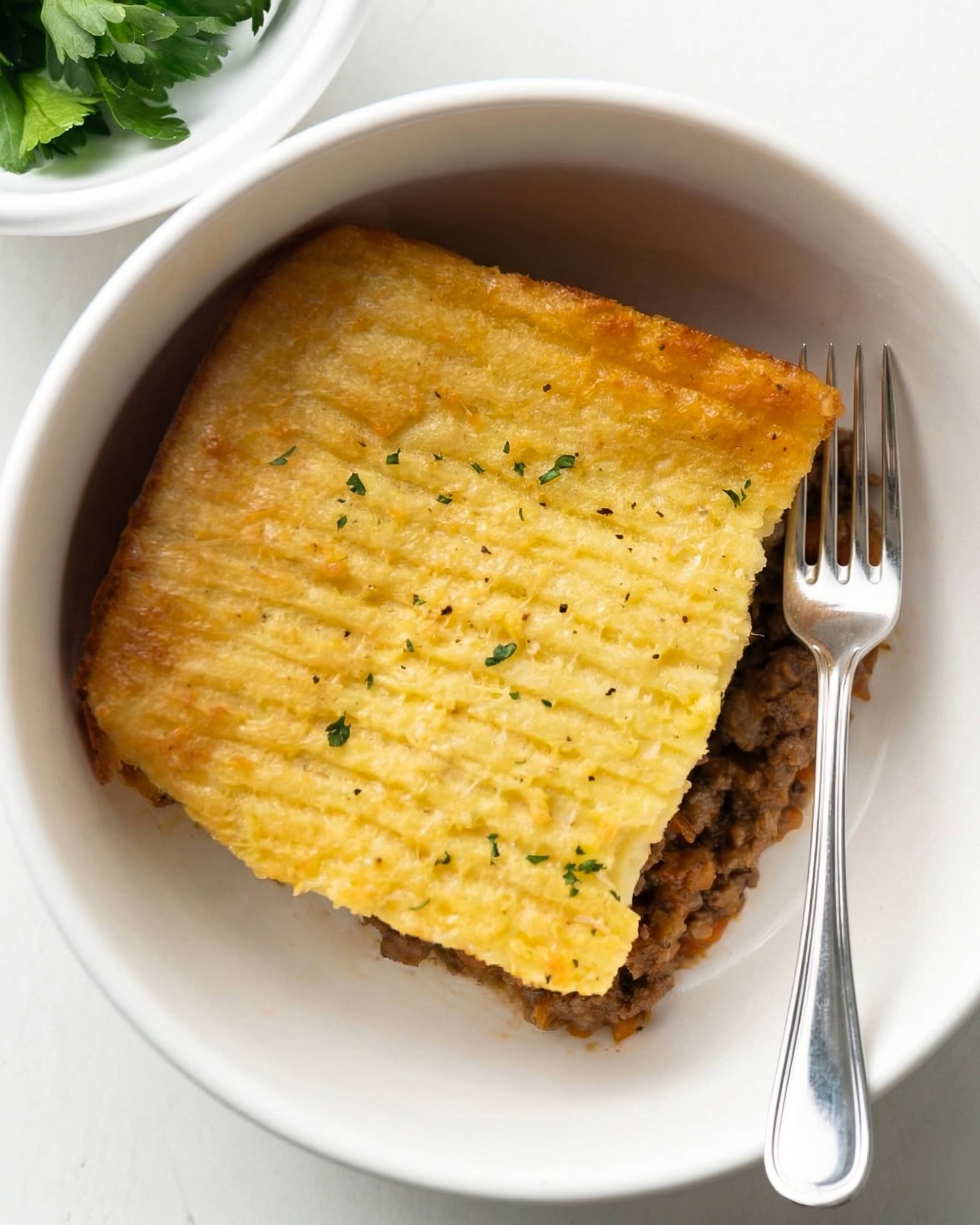 A close-up, overhead view of a single square serving of gluten-free Shepherd’s Pie in a white bowl. The serving features a rich ground lamb filling topped with a golden-brown, textured mashed potato crust and fresh parsley. A silver fork rests beside the pie in the bowl.