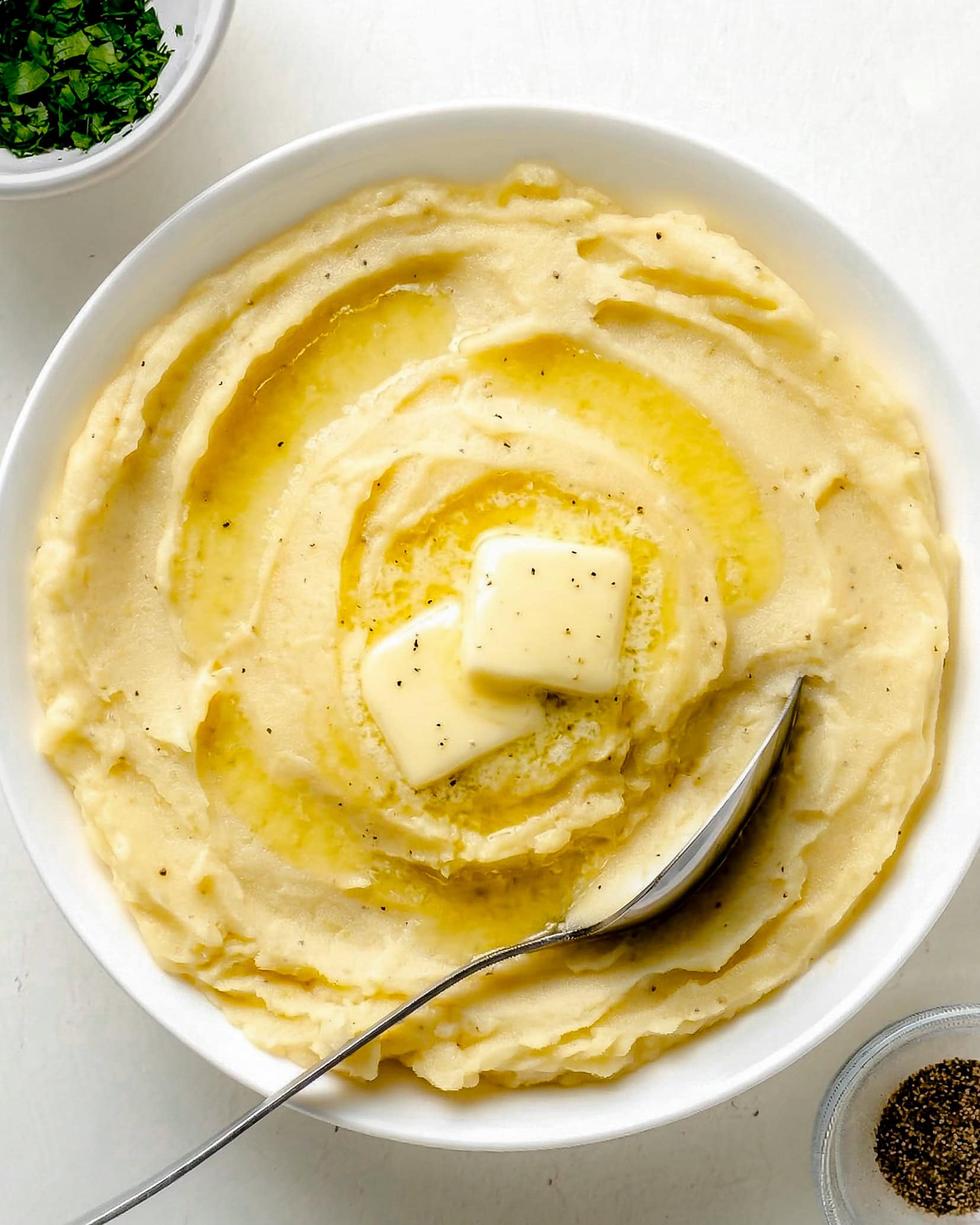 Mashed Yukon Gold Potatoes in a white serving bowl with two pats of butter in the center, and a bowl or parsley and black pepper off the the sides.