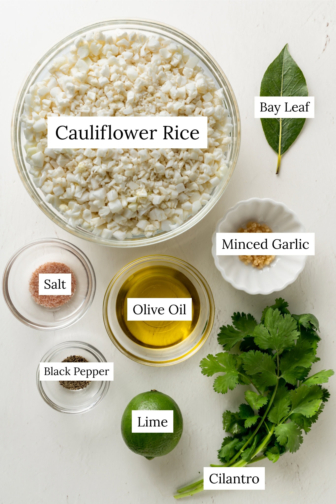 Ingredients for cilantro lime cauliflower rice. A bowl of cauliflower rice, bay leaf salt, black pepper, olive oil. minced garlic, cilantro, and one lime.