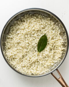Cauliflower rice with a bay leaf in a large stainless steel pan.