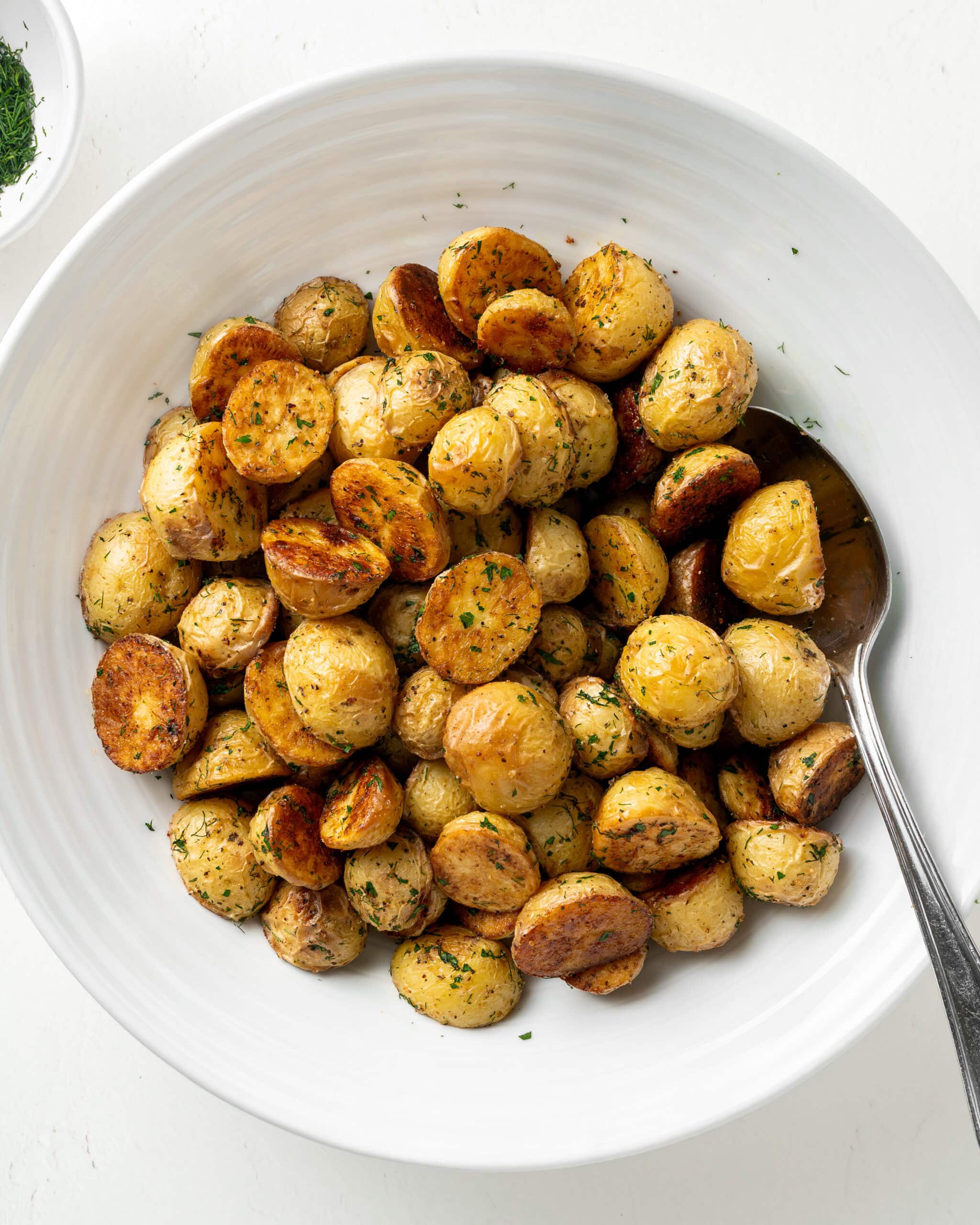 Garlic Dill Roasted Potatoes in a Large white Serving Bowl with a silver spoon. Across from the bowl is a small bowl of chopped dill.