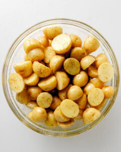 Baby yellow potatoes in a large clear bowl, cut in half with added oil and spices coated.